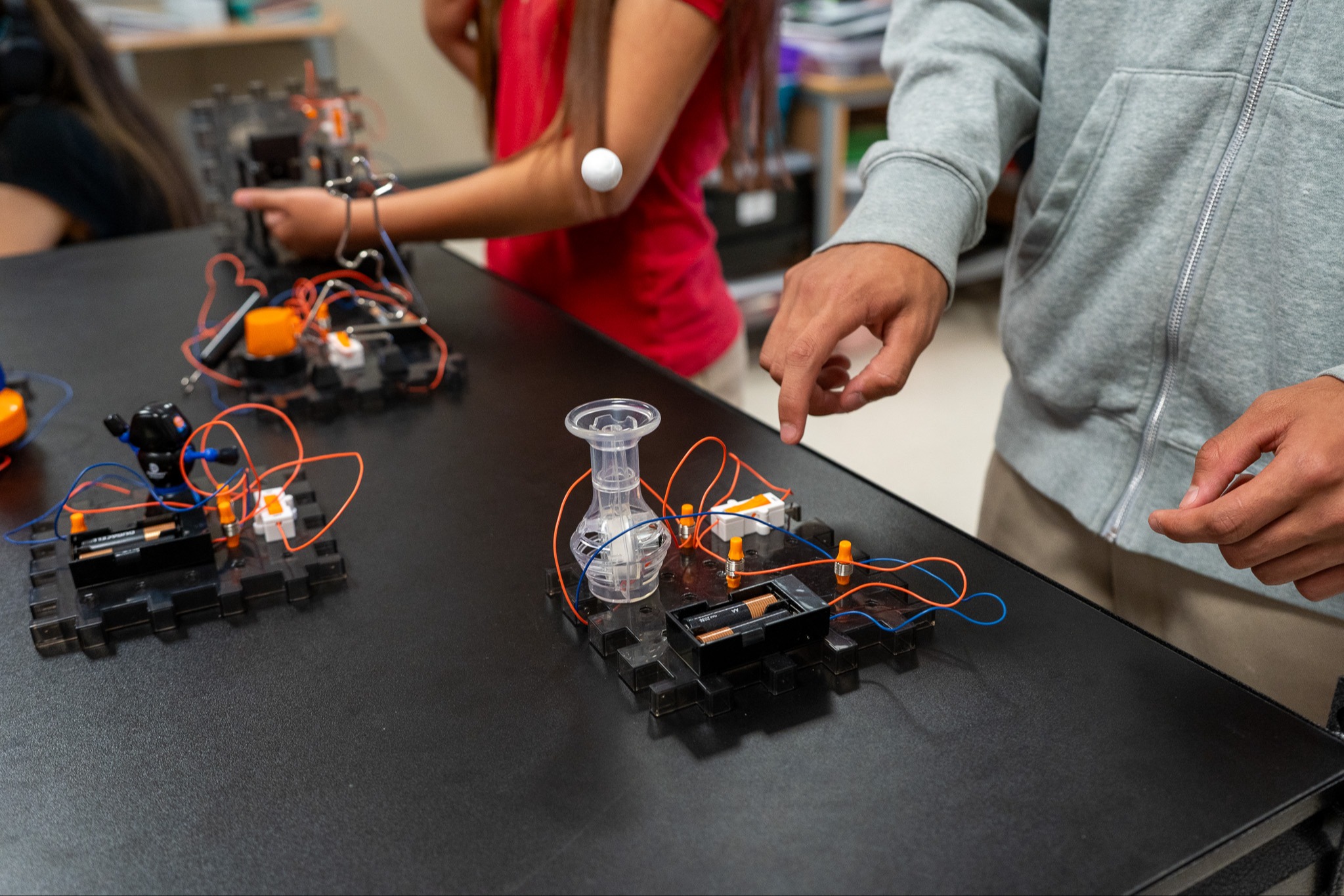 Two people in a lab with electronic circuits and robotic parts on a table. One person points to a device.