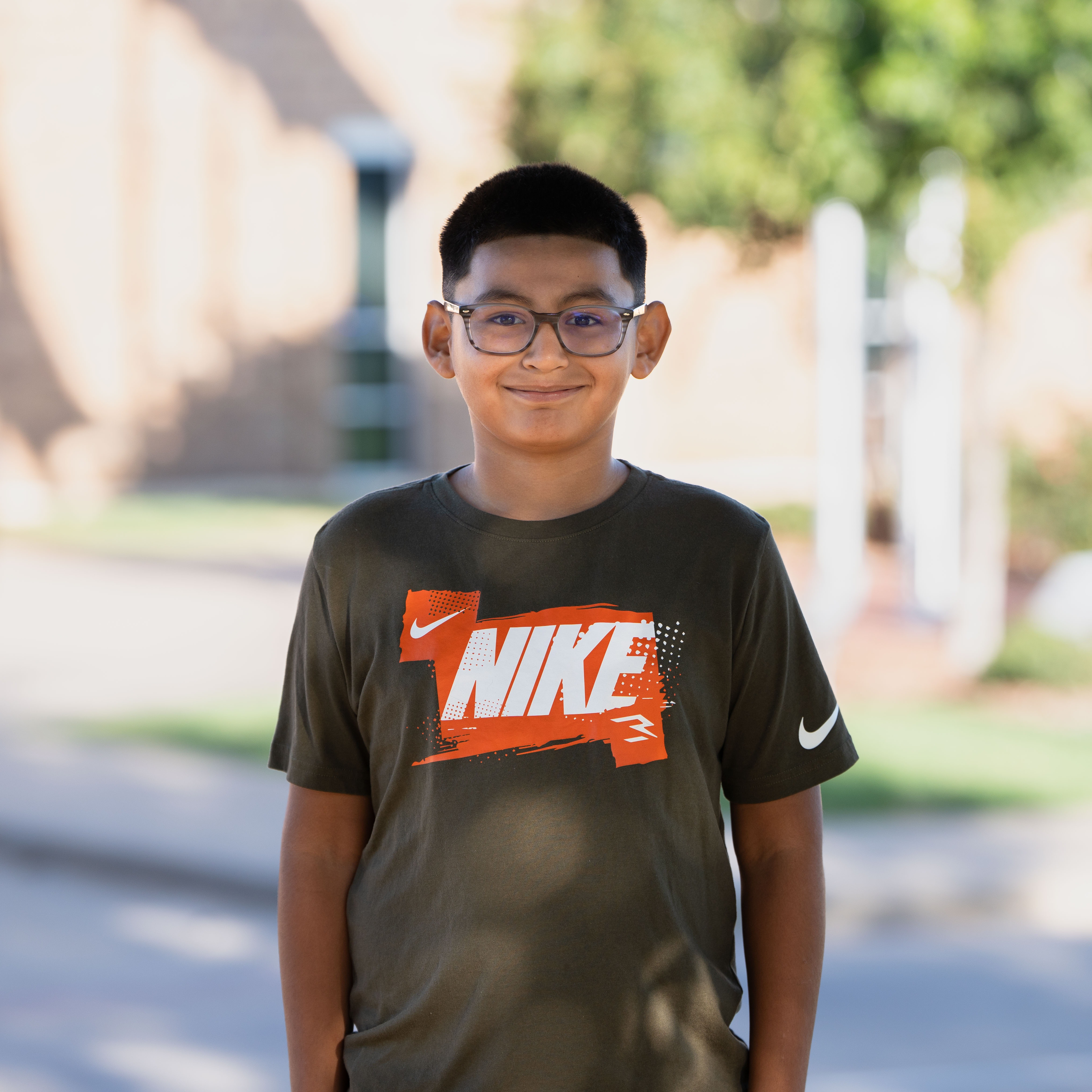 A boy wearing glasses and a black Nike shirt stands on a road in front of a building.