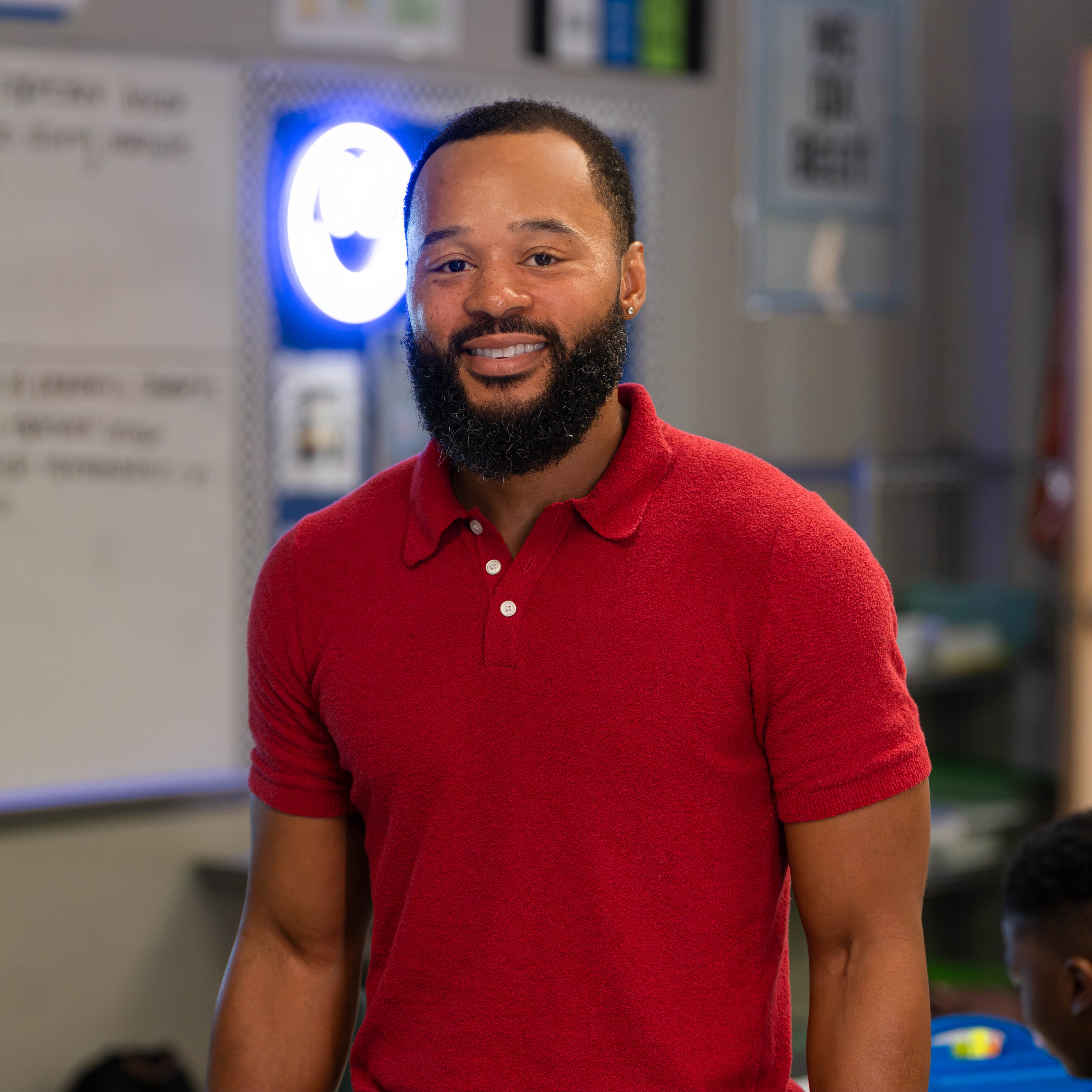 A man with a beard wearing a red polo stands in front of a whiteboard in a classroom.