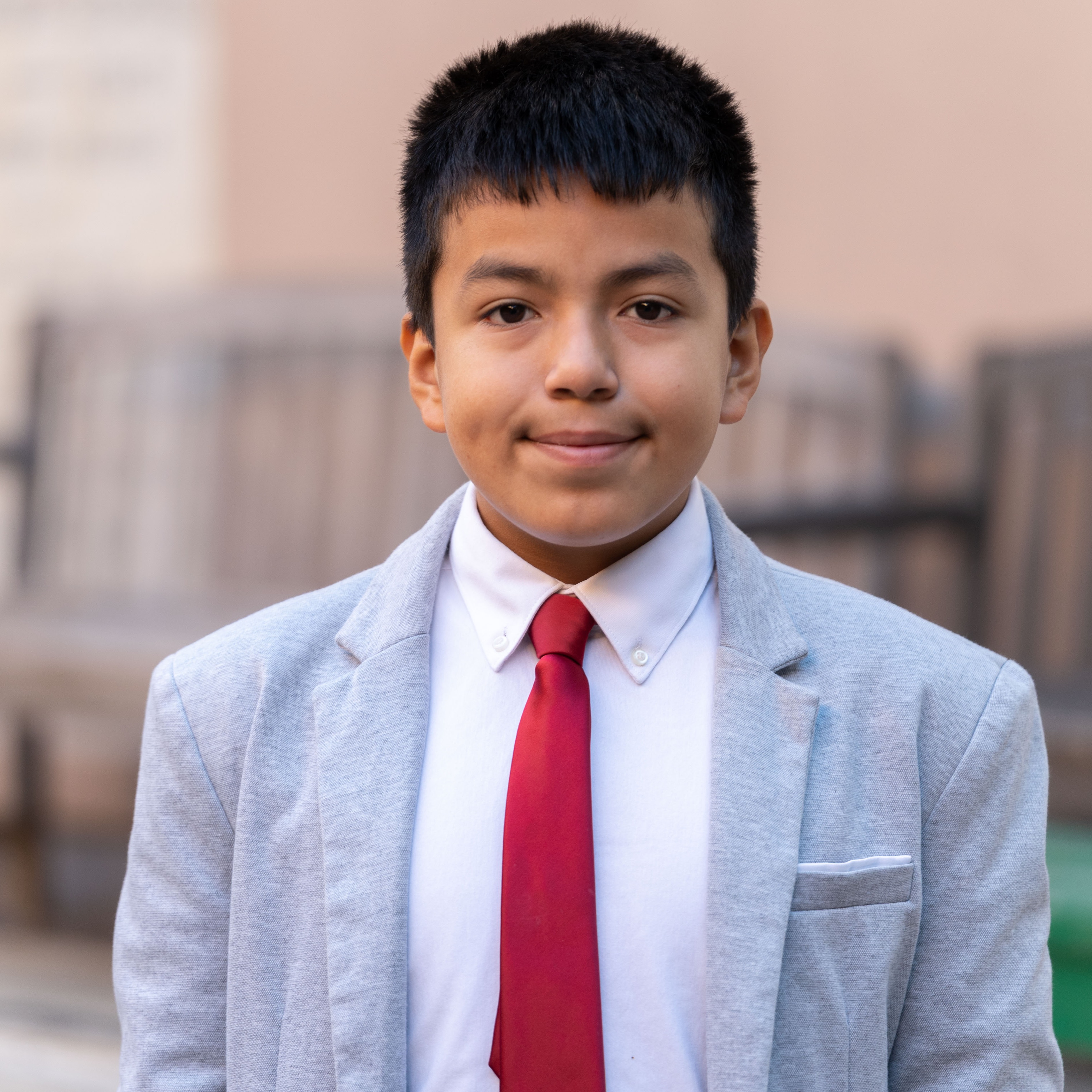 Boy in a gray jacket and red tie stands against a background of wooden benches.