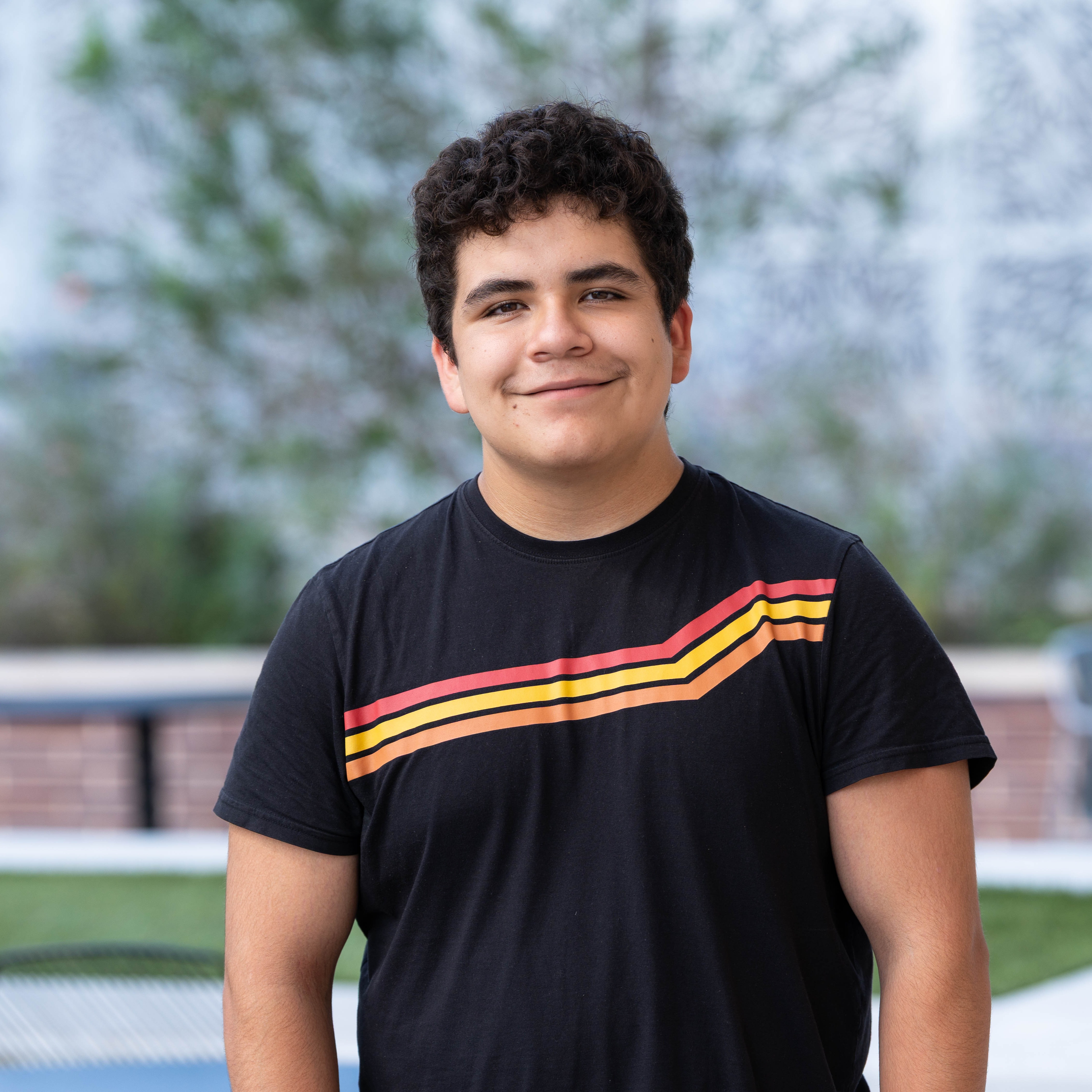 A young person in a black t-shirt with a multicolored stripe stands outside, smiling at the camera. Behind them is a grassy area and a brick wall.