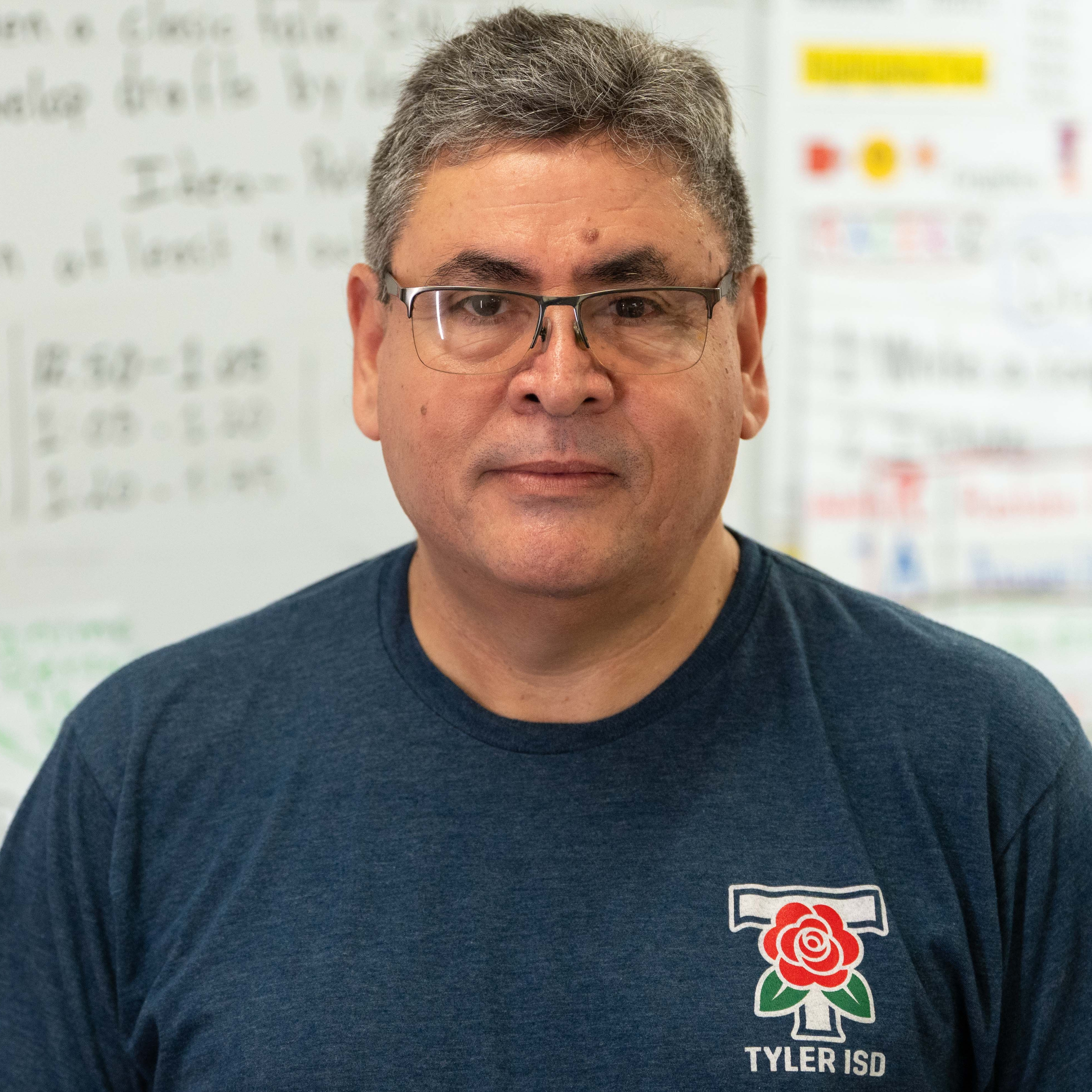 Person standing in front of whiteboard. Person wears glasses and a navy blue shirt with a logo.