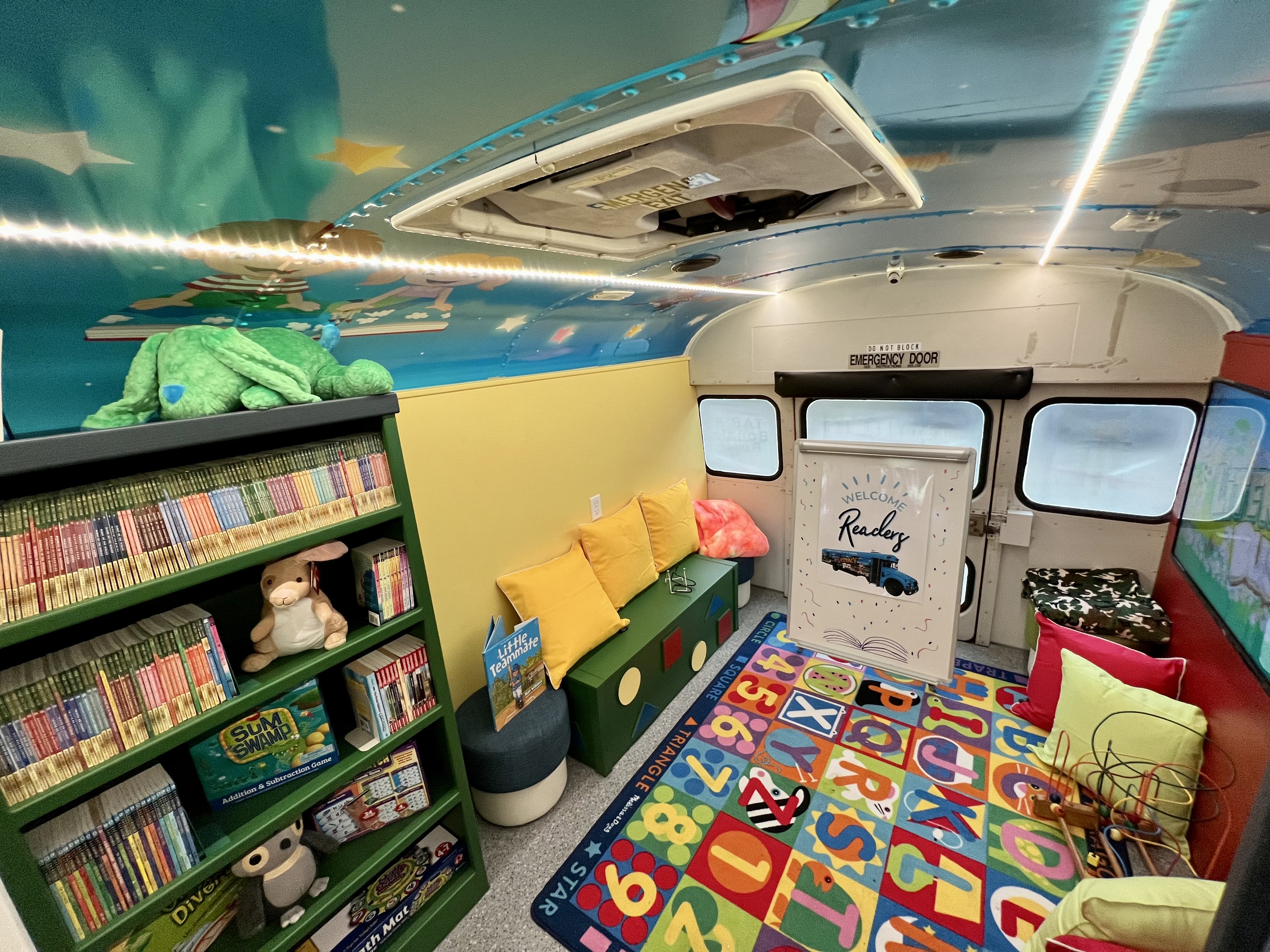 inside of school bus painted in bright colors on roof. All seats have been removed and bookshelves lined with books are along the walls. Chairs, cushions, stuffed animals are also inside