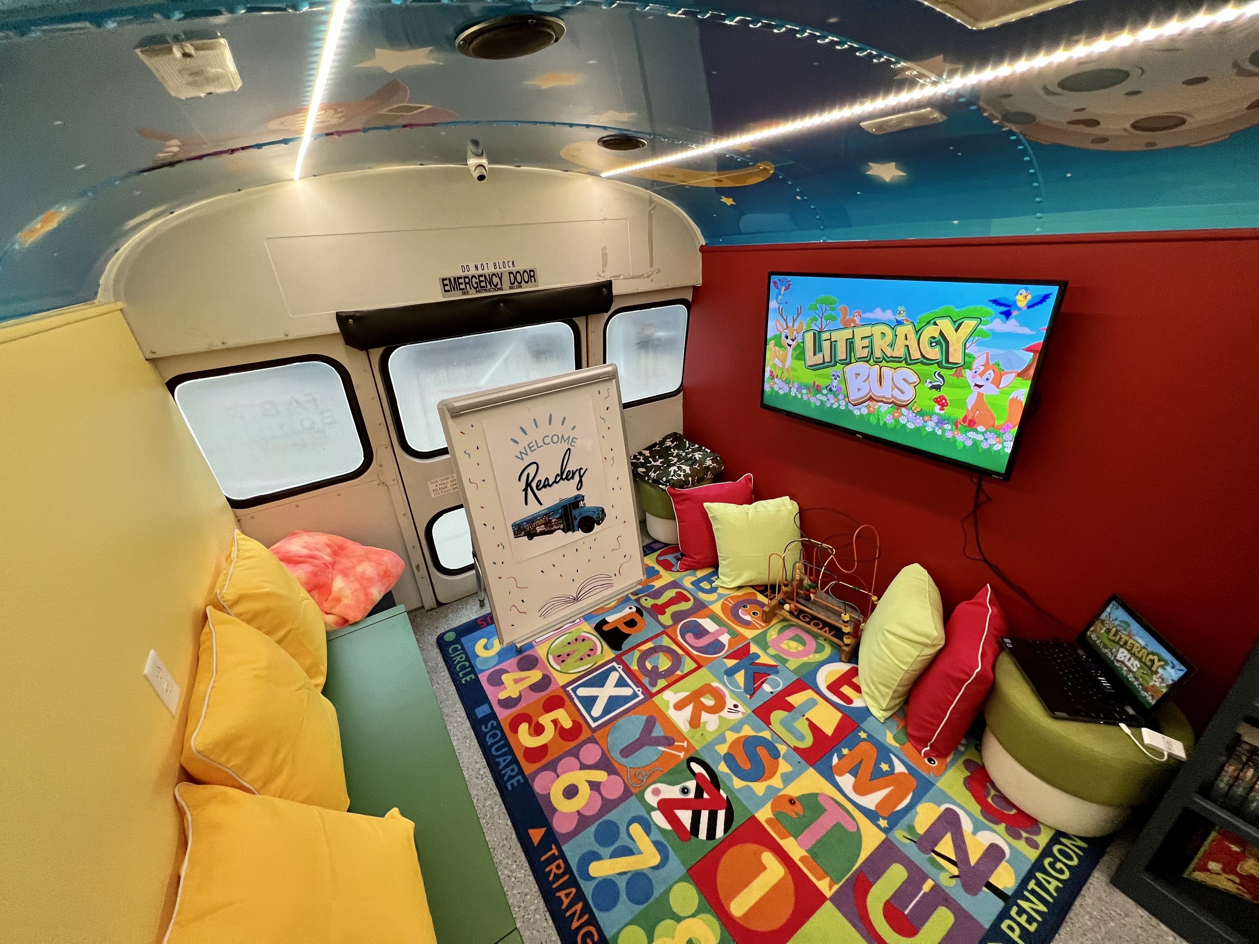 inside of school bus painted in bright colors on roof. All seats have been removed and bookshelves lined with books are along the walls. Chairs, cushions, stuffed animals are also inside