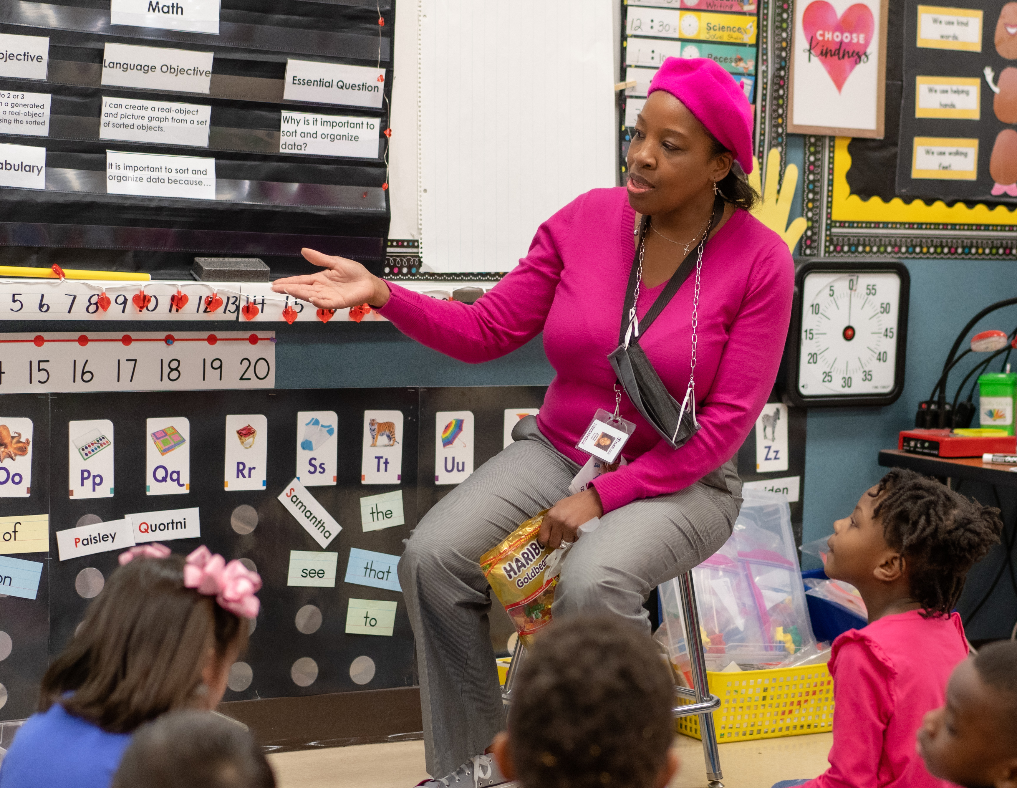 African American teacher wearing hot pink shirt and head covering sits on stool in front of her young students