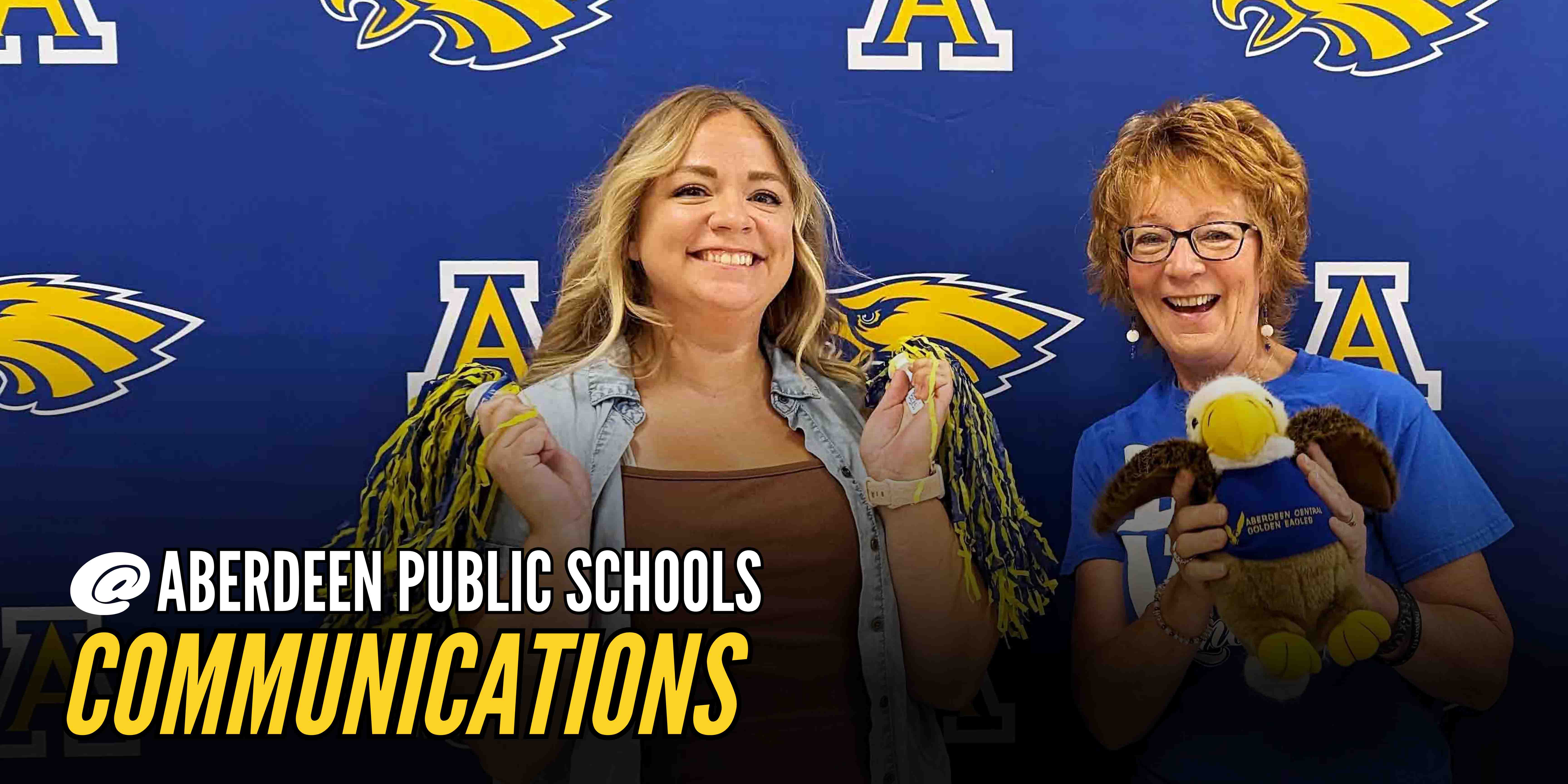 Two staff members holding Aberdeen Public Schools gear in front of the backdrop with text: @ Aberdeen Public Schools Communications