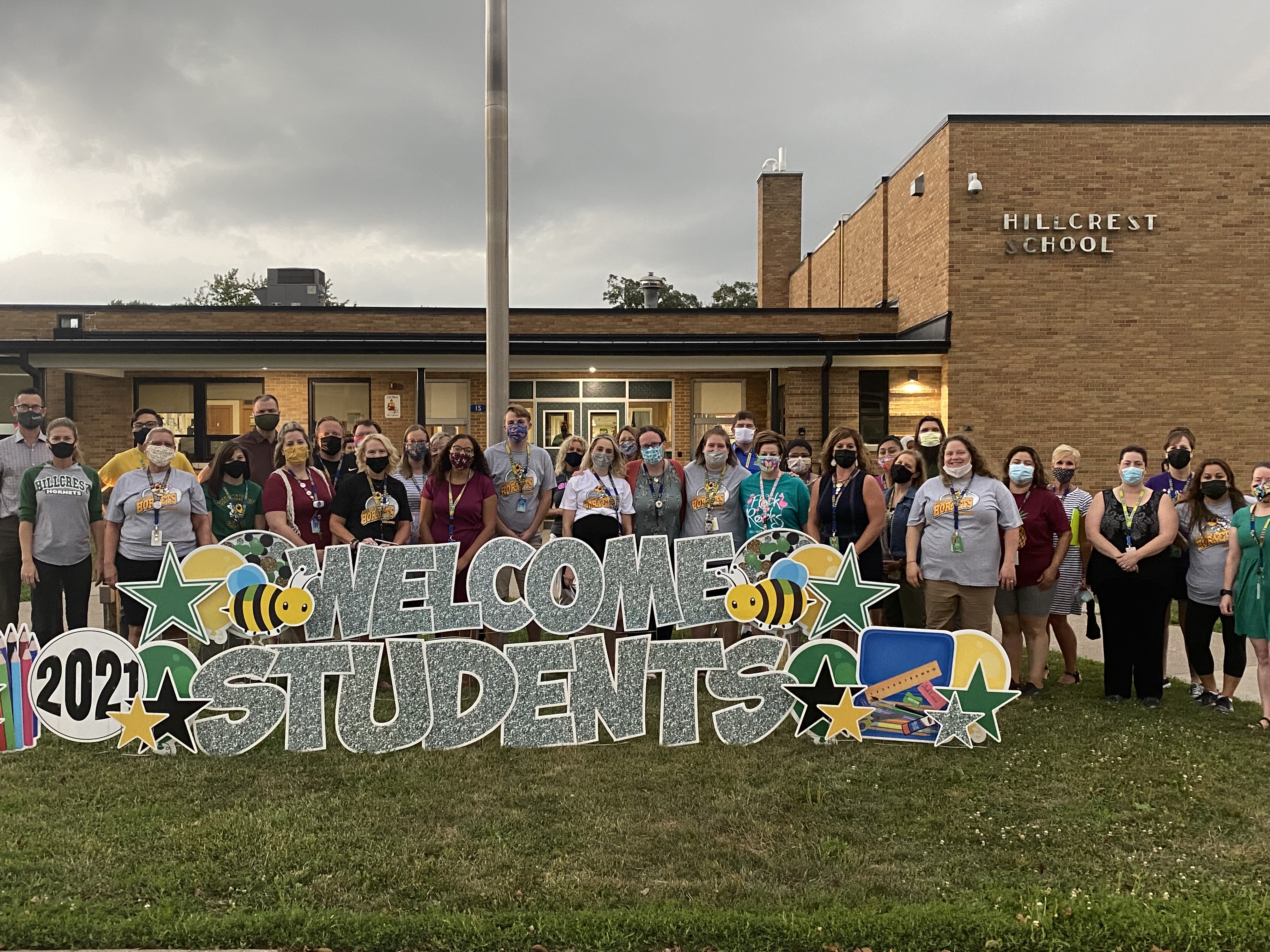 Hillcrest staff on the first day of school. They are standing outside in front of the building. Large signs that say Welcome Back Students are stuck in the grass and the teachers smile while standing behind the signs.