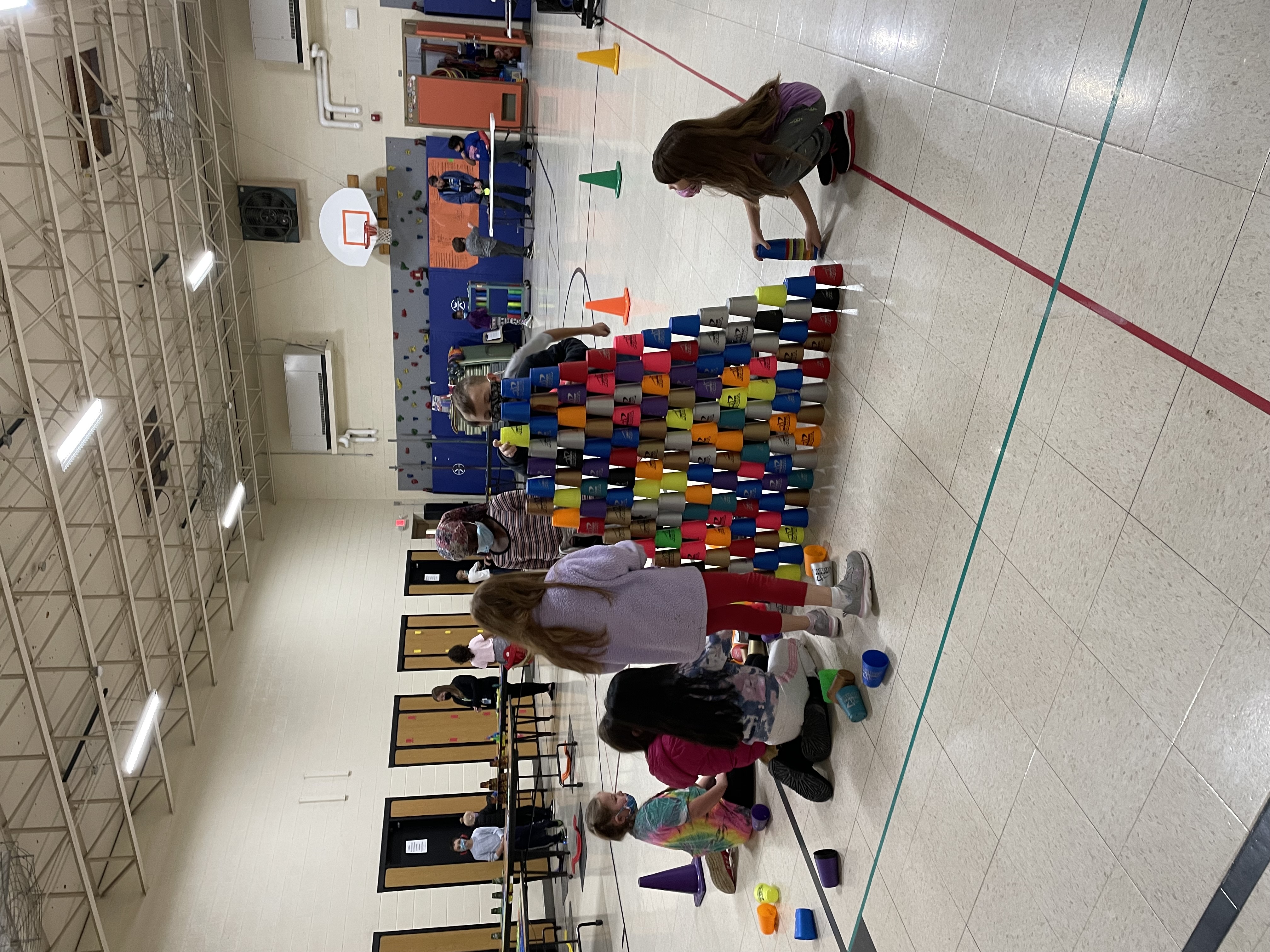 a group of children working together to stack and  create a large cup tower. The cup tower is on the floor and is eye level with the children standing next to it.
