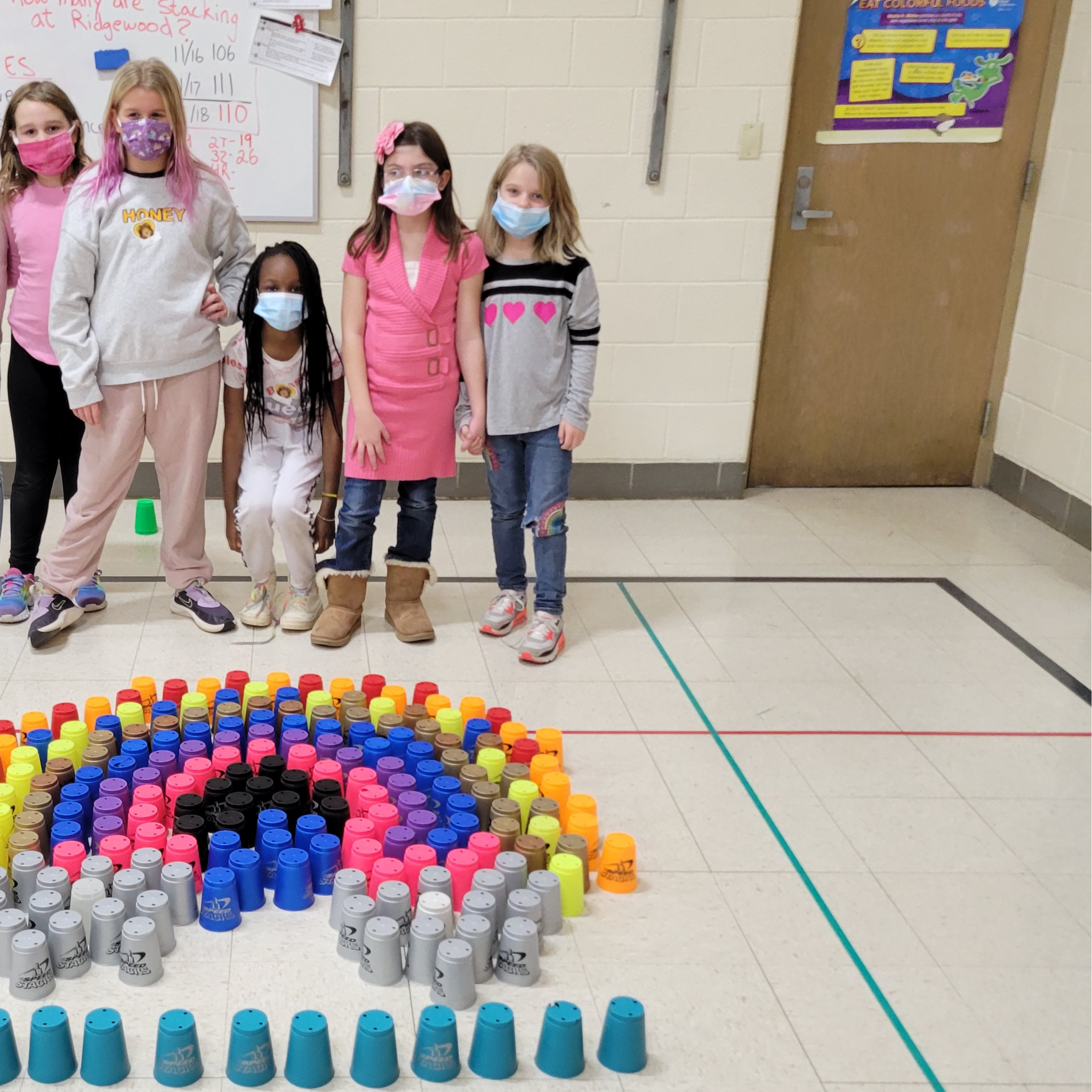 a group of girls stand behind a rainbow they made out of different colored  cups on the floor.  At the bottom of the rainbow the students added gray cups so it looks like clouds under the rainbow