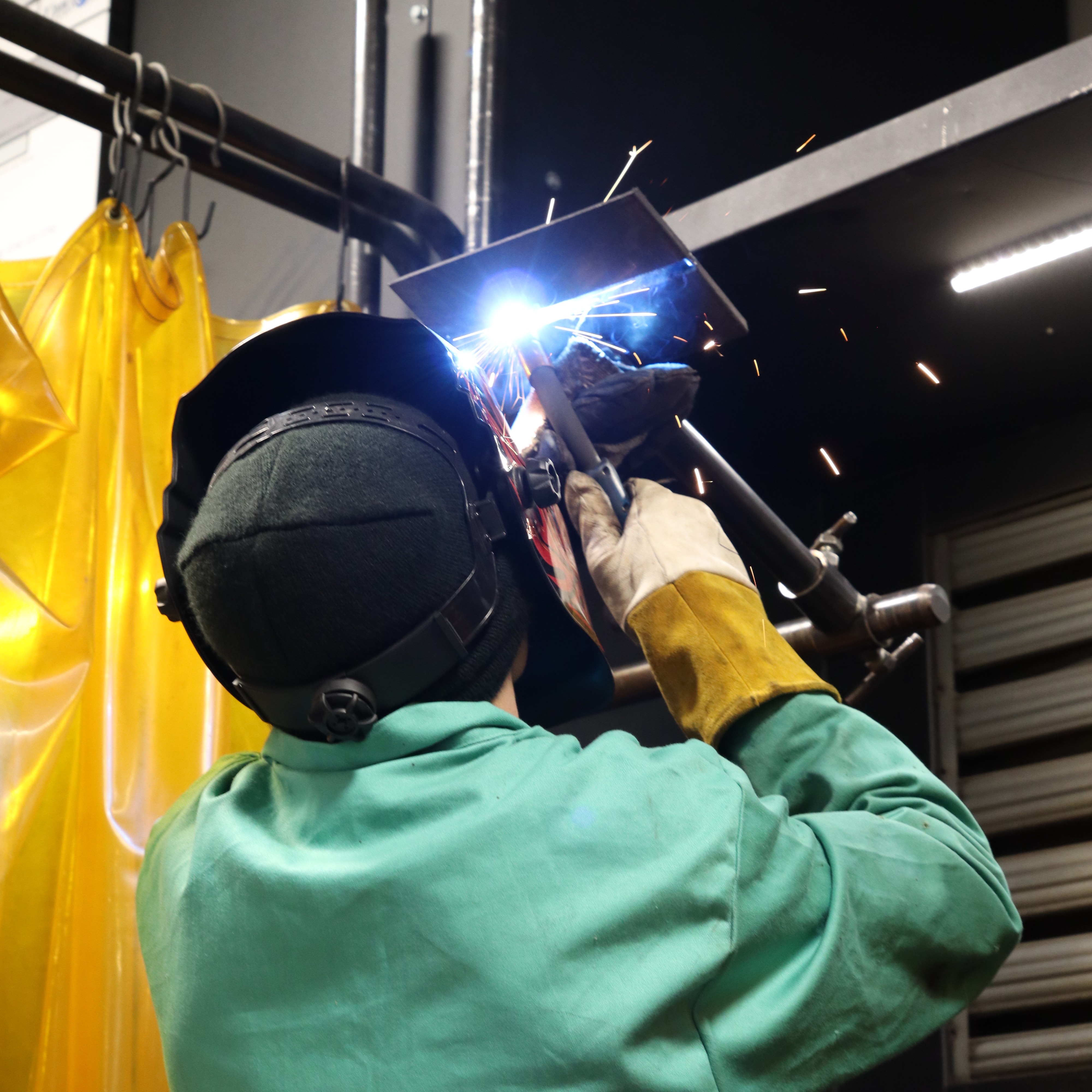 Person in a green jacket and protective helmet welding metal with a torch. Yellow protective sheet on the left.