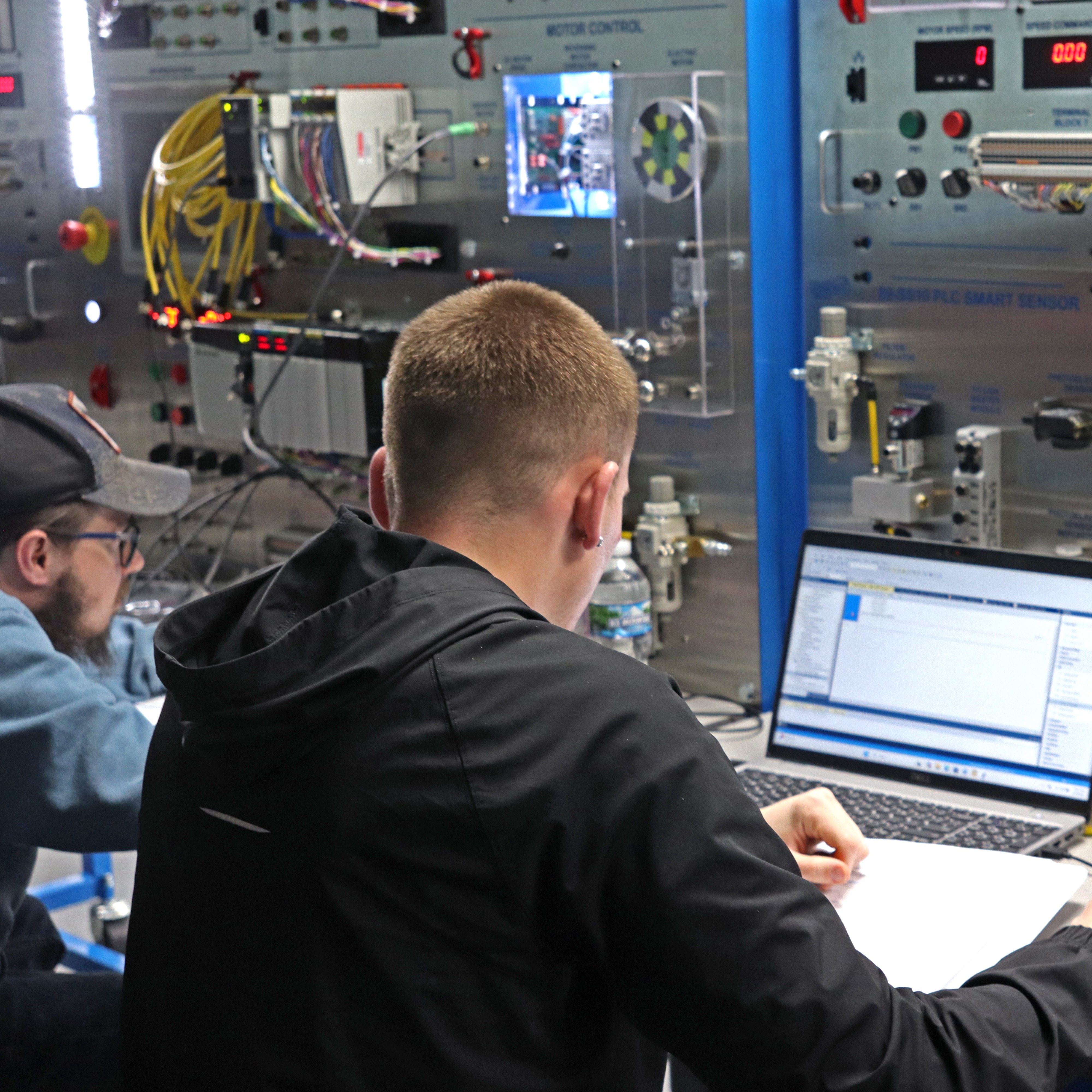 A man wearing a black jacket sits at a desk in front of a computer. Behind him, complex machinery is visible.