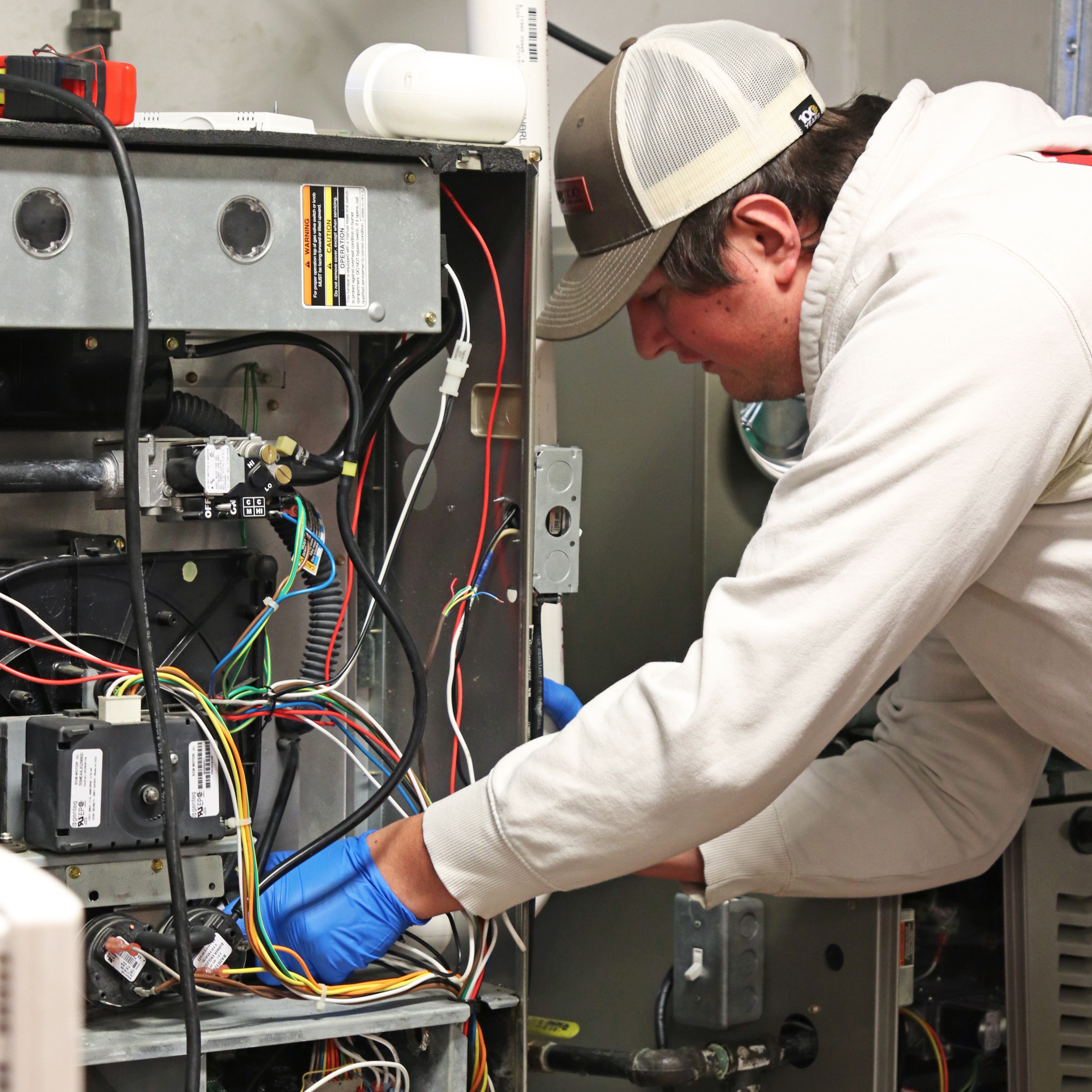 Technician in blue gloves and a cap working on a heating unit with exposed wiring.