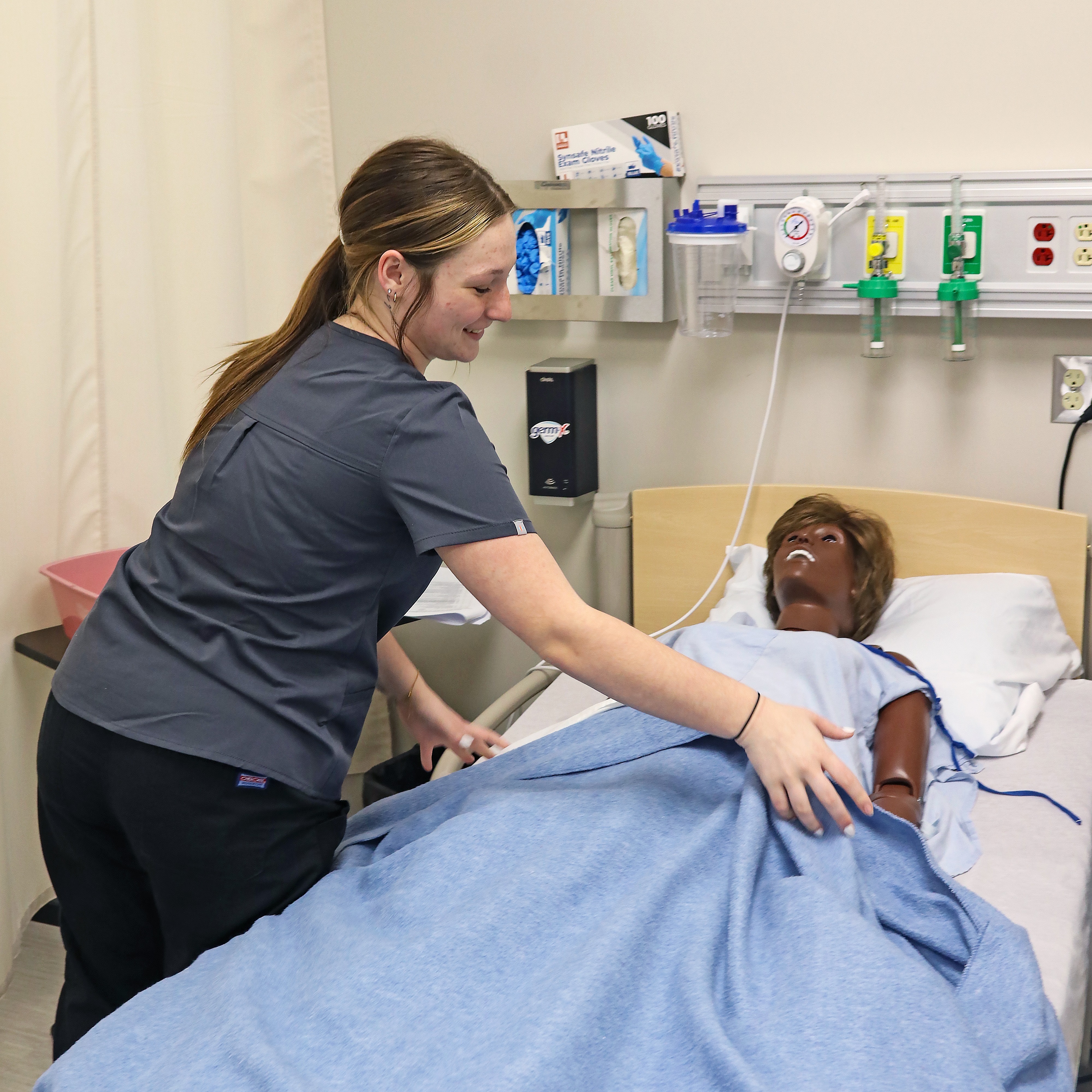 A woman in scrubs bends over a hospital bed, holding the hand of a mannequin wearing a hospital gown.