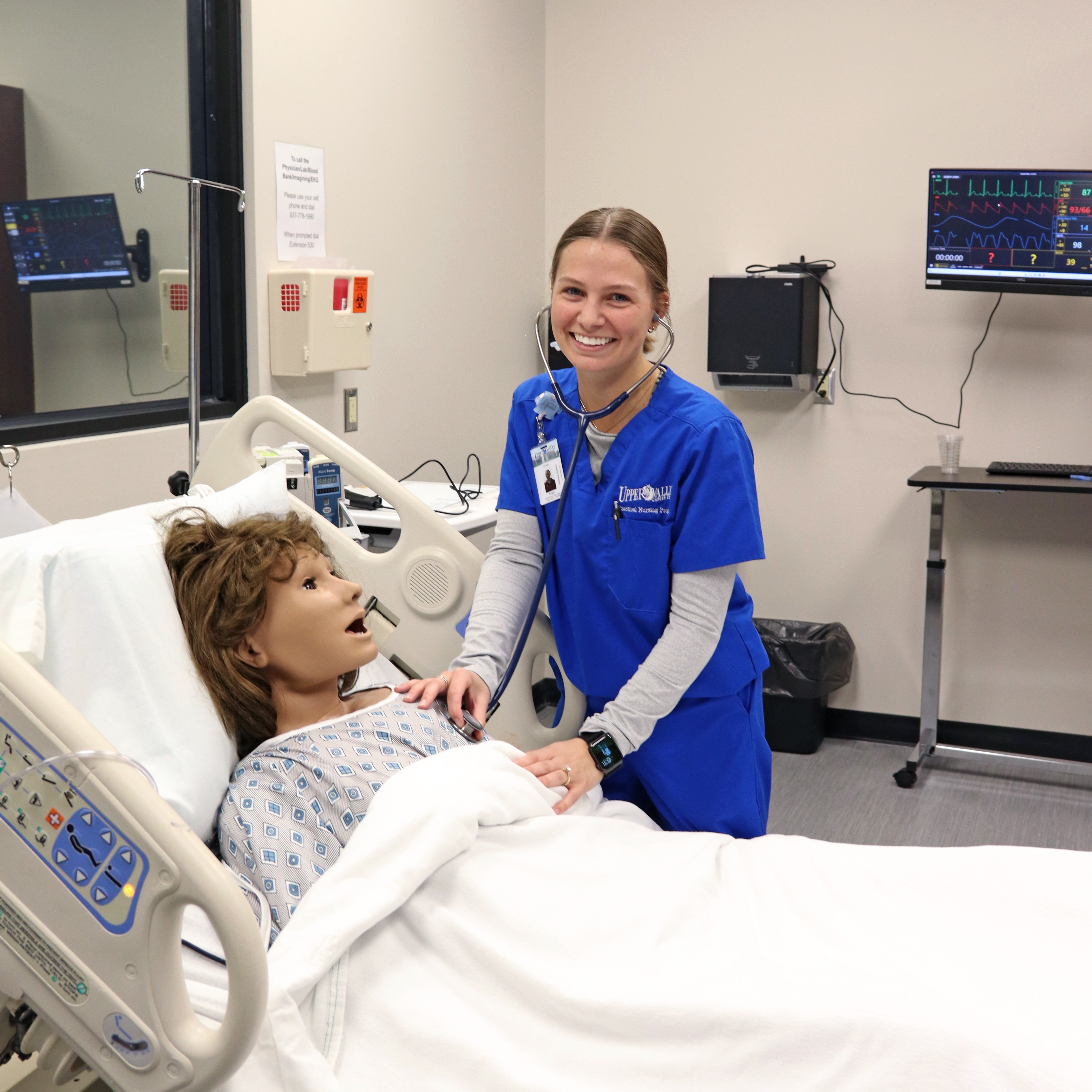 A woman in blue scrubs stands beside a hospital bed with a mannequin, smiling. The room has medical monitors and equipment.