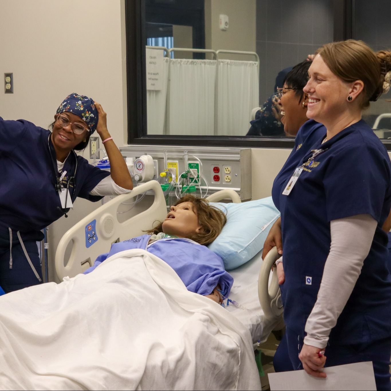 Hospital room with three healthcare workers. A mannequin lies on the bed, and a nurse stands by the window.