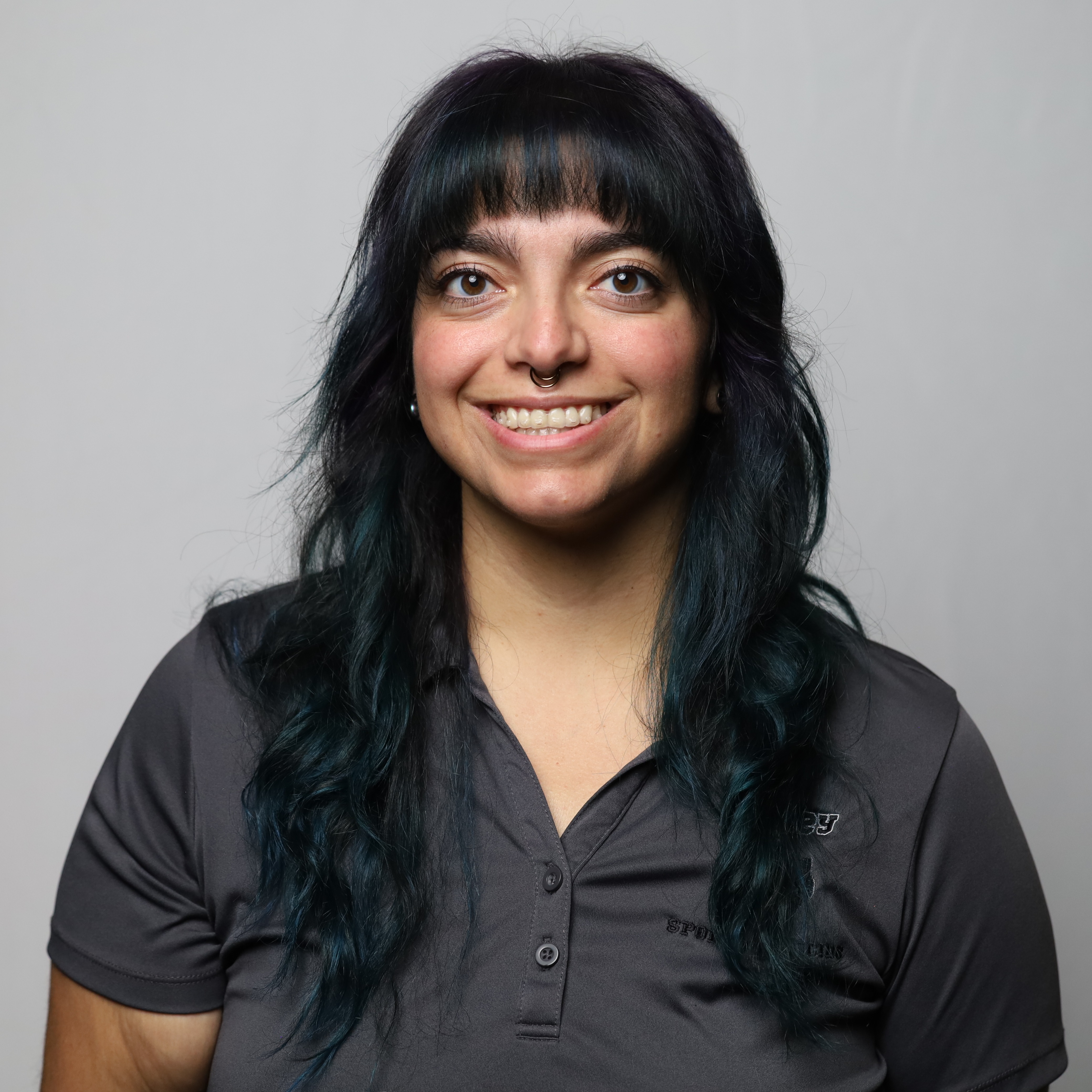 Woman with black hair wearing a gray polo. She is standing and smiling, with gray background.