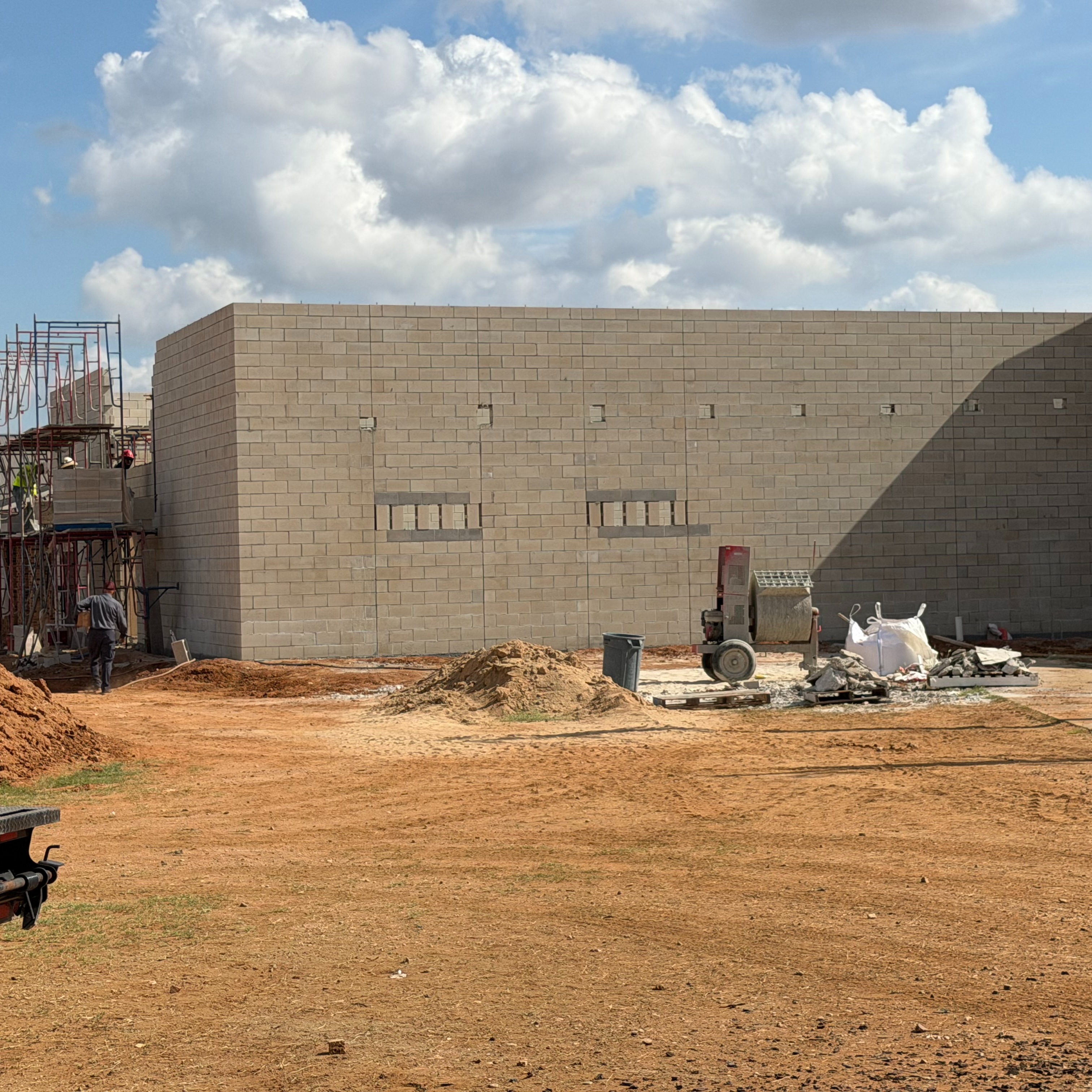An under-construction building with a large wall, windows, and a dirt ground. A vehicle is parked on the left.