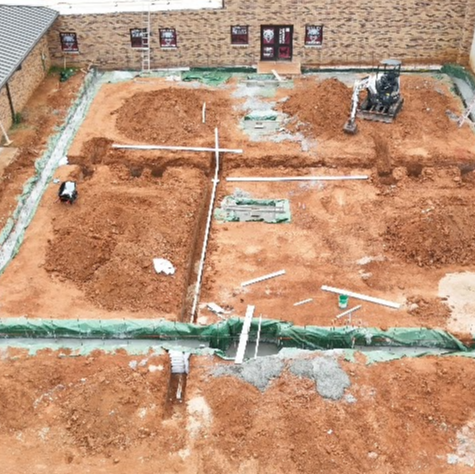 Aerial view of a construction site with a building, dirt, and a square-shaped foundation surrounded by green strips.
