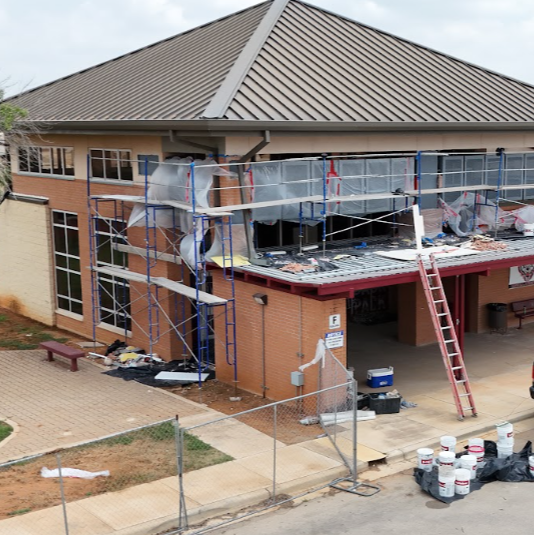 A building under construction. Wooden scaffolding covers a portion of the roof. White paint buckets and tarps are scattered around.