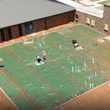 A top view of a construction site with workers laying green tiles. Two buildings with gray roofs are partially visible.