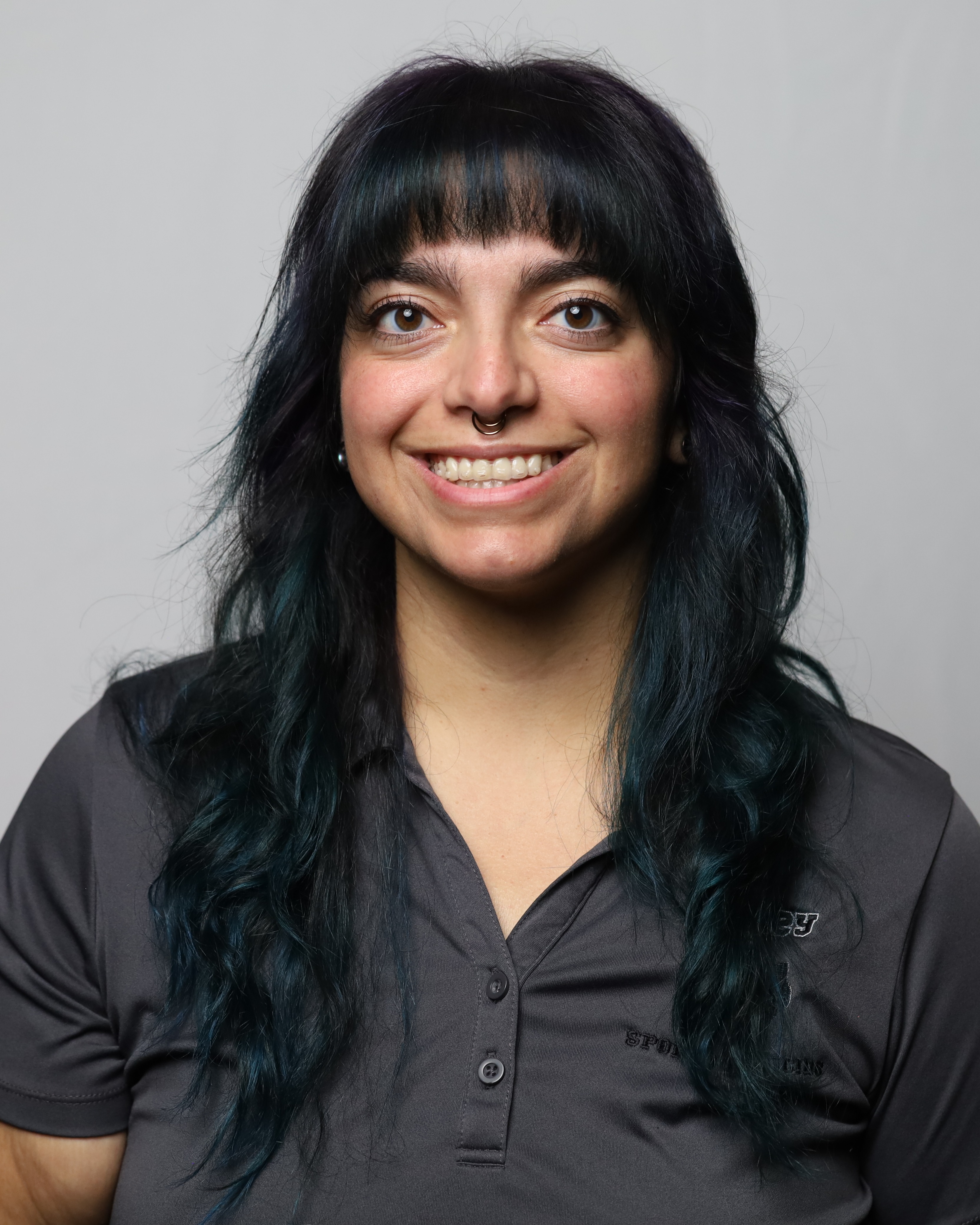 A woman with long black hair, smiling and looking at the camera, stands against a plain gray background.