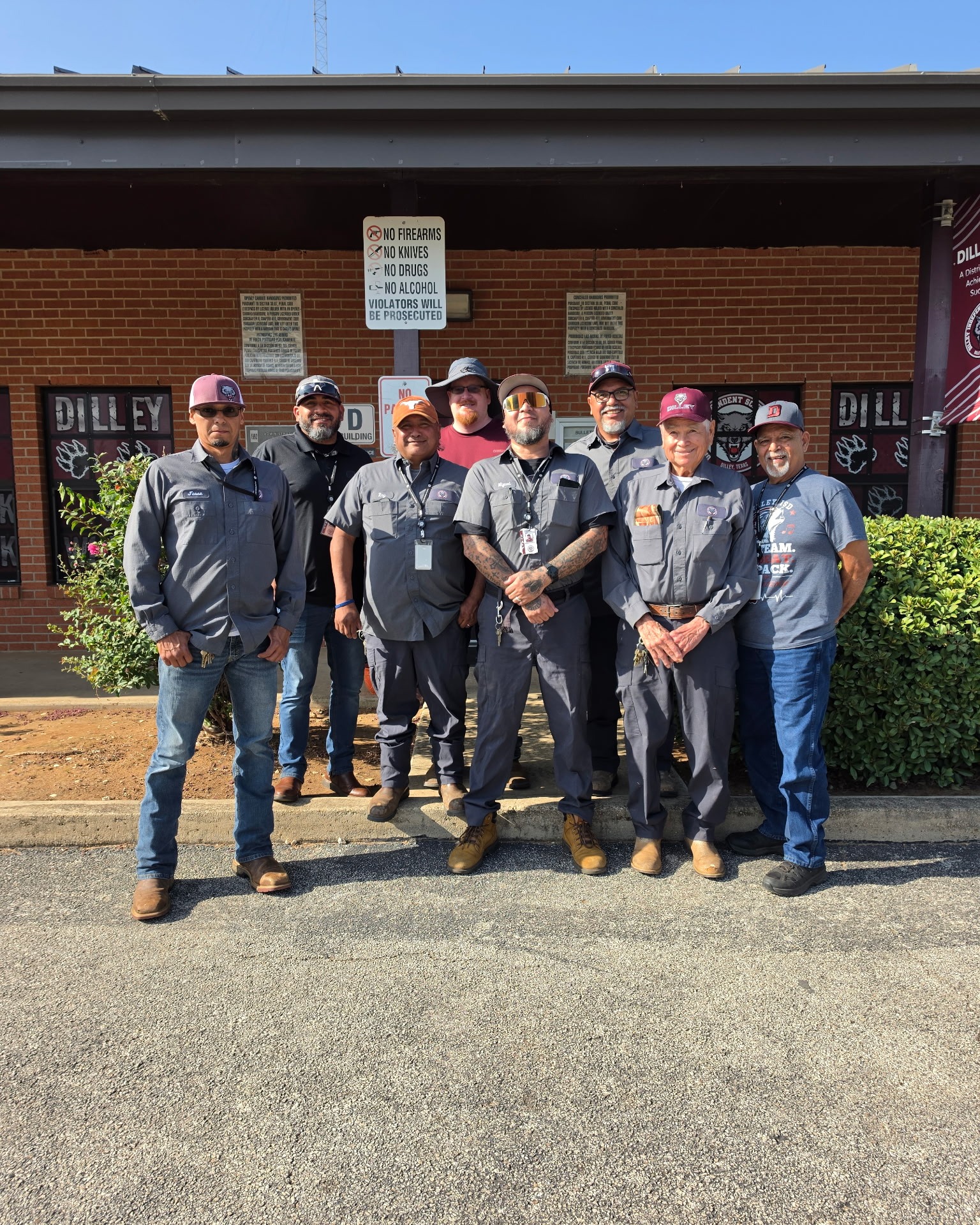 Eight people in uniforms and hats, some with glasses, pose for a photo in front of a building.