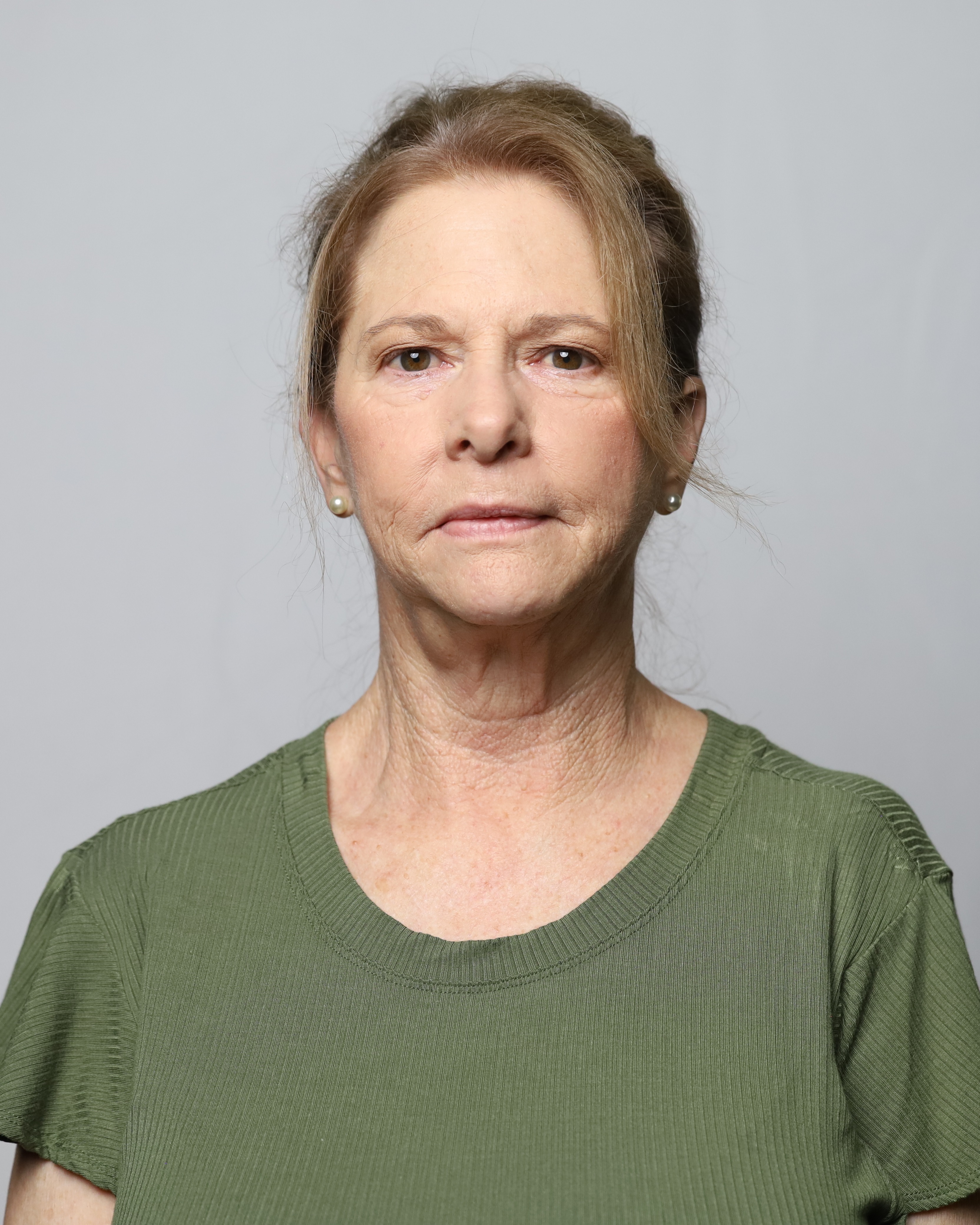 An older woman stands against a gray background, wearing a green shirt. She has short hair and a serious expression.