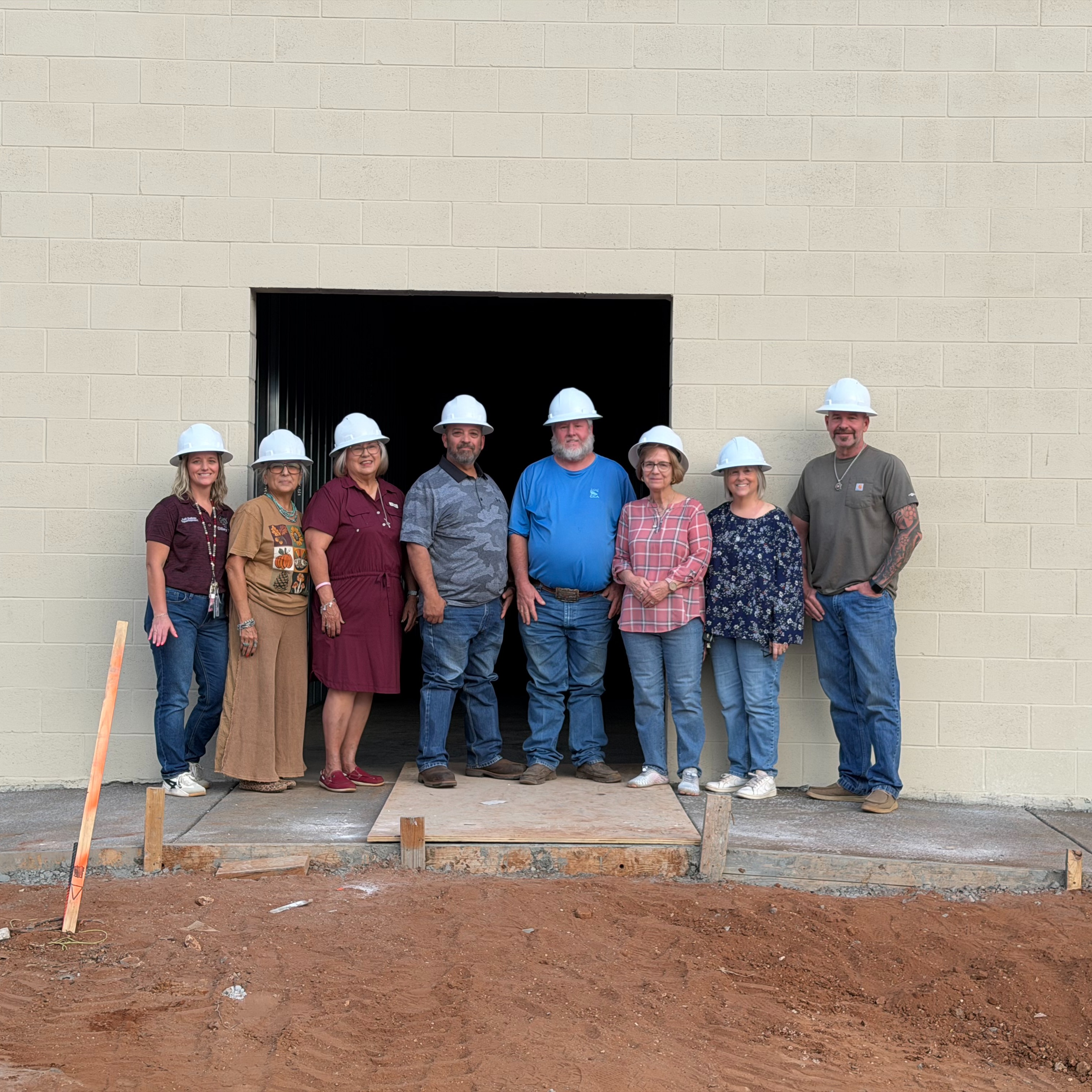 Group of adults in white hard hats standing outside a building under construction. Some are on a concrete slab.