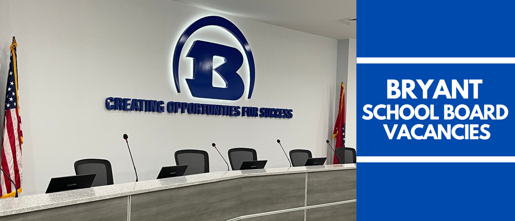Conference room with long table, multiple chairs, and microphones. Wall has logo, text, and flags.