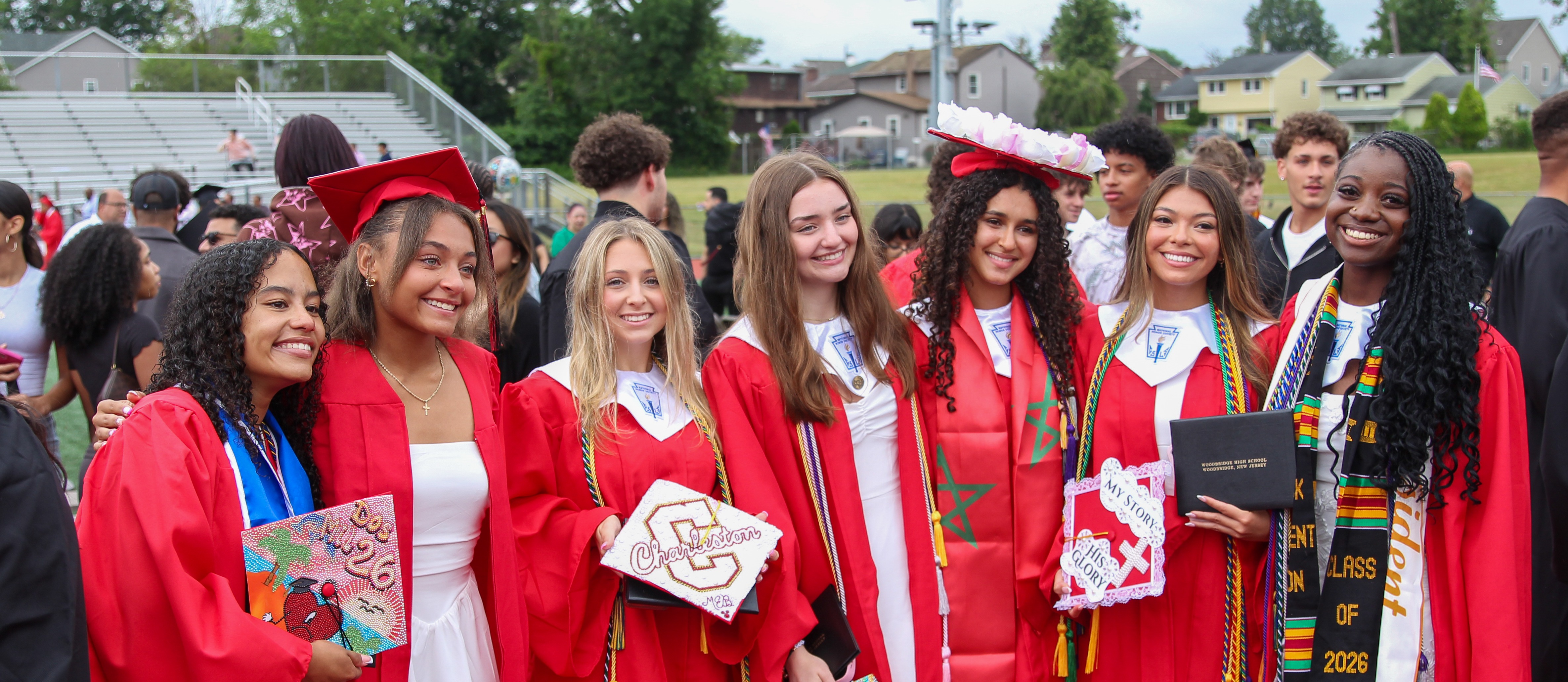 A group of women in red graduation gowns and hats stand together on a sports field.