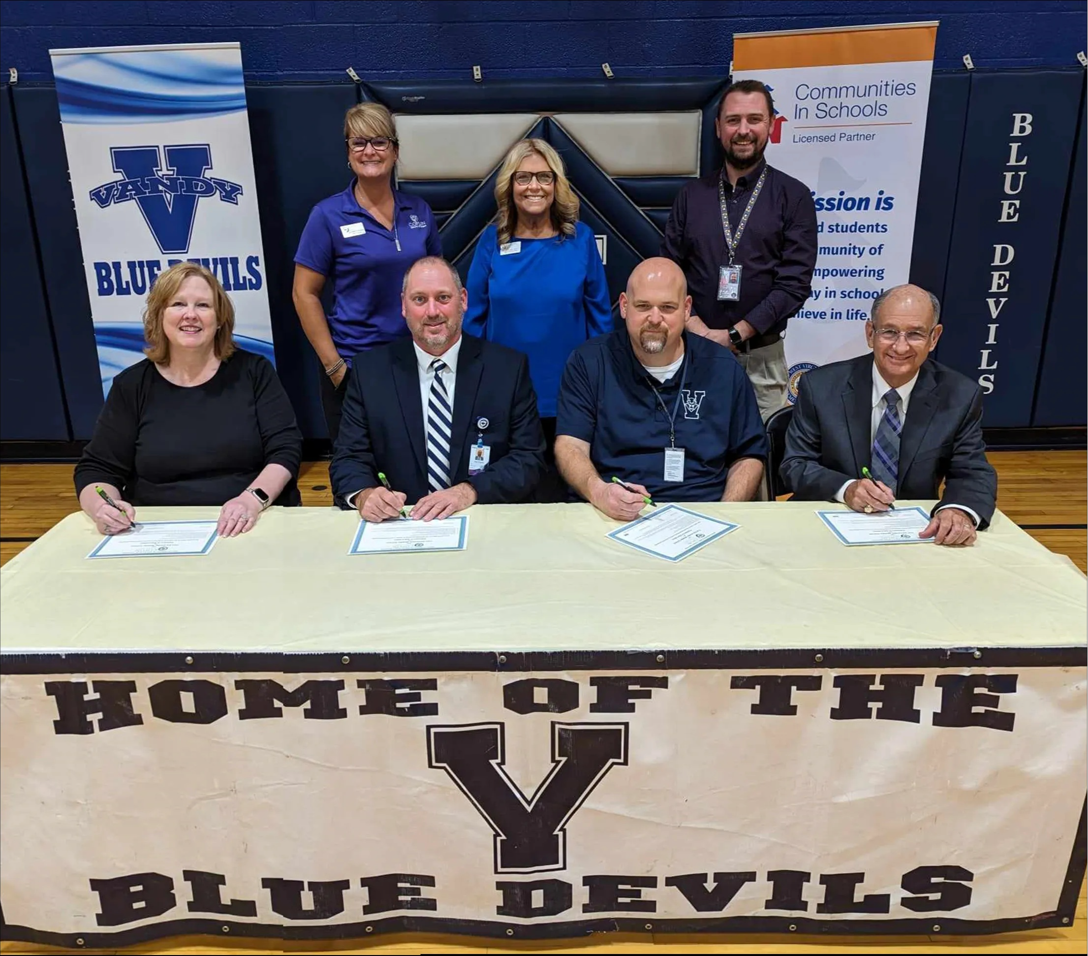 members of the new education - business partnership sitting at a table and standing in a row behind inside a school gymnasium