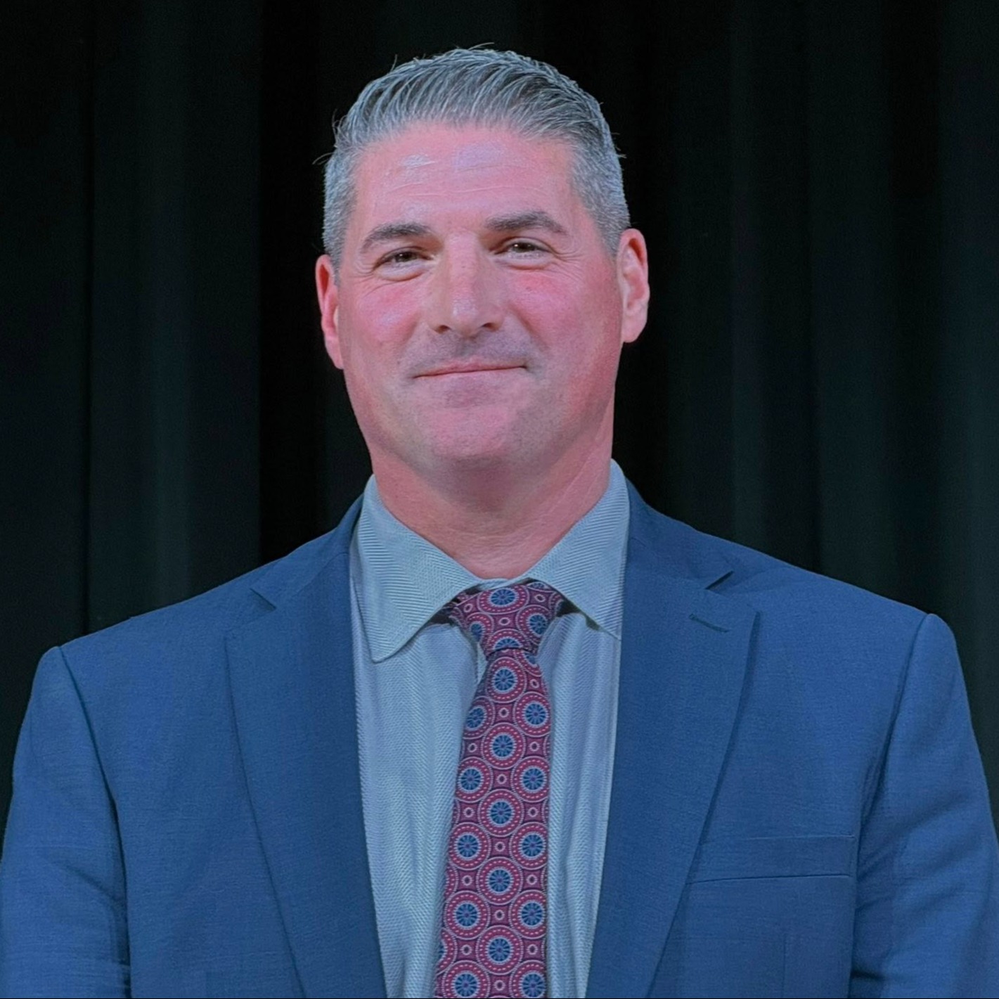 A man in a navy blue suit and tie, smiling, standing before a black backdrop.
