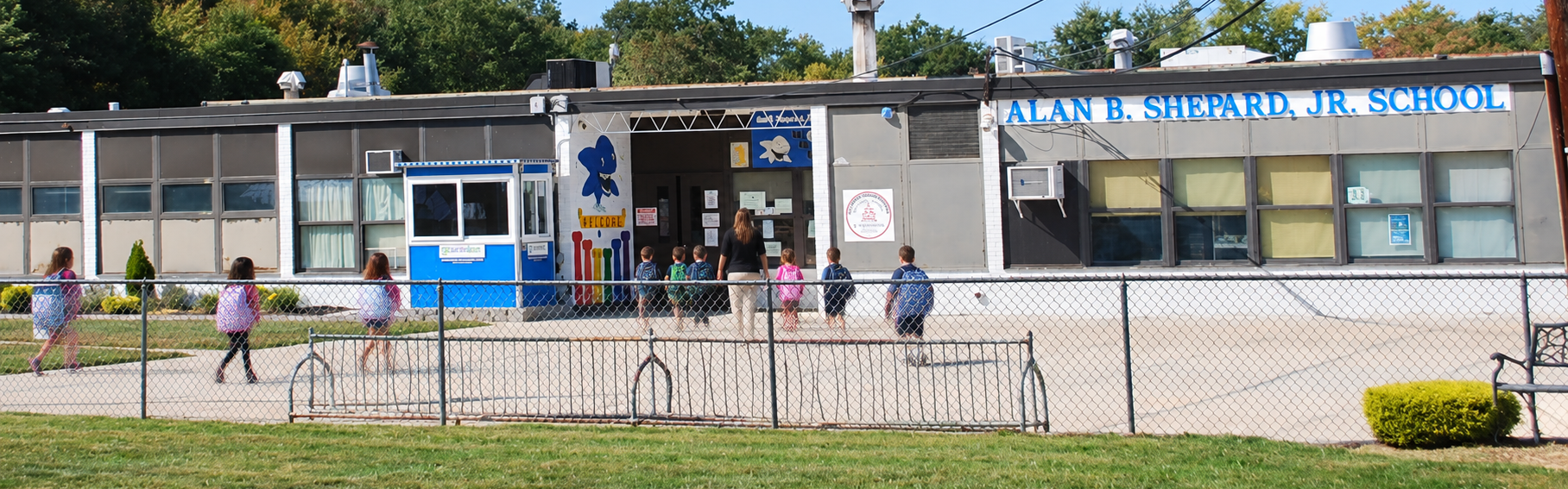 Exterior of a school with children walking to the entrance. A grassy area with a metal fence.