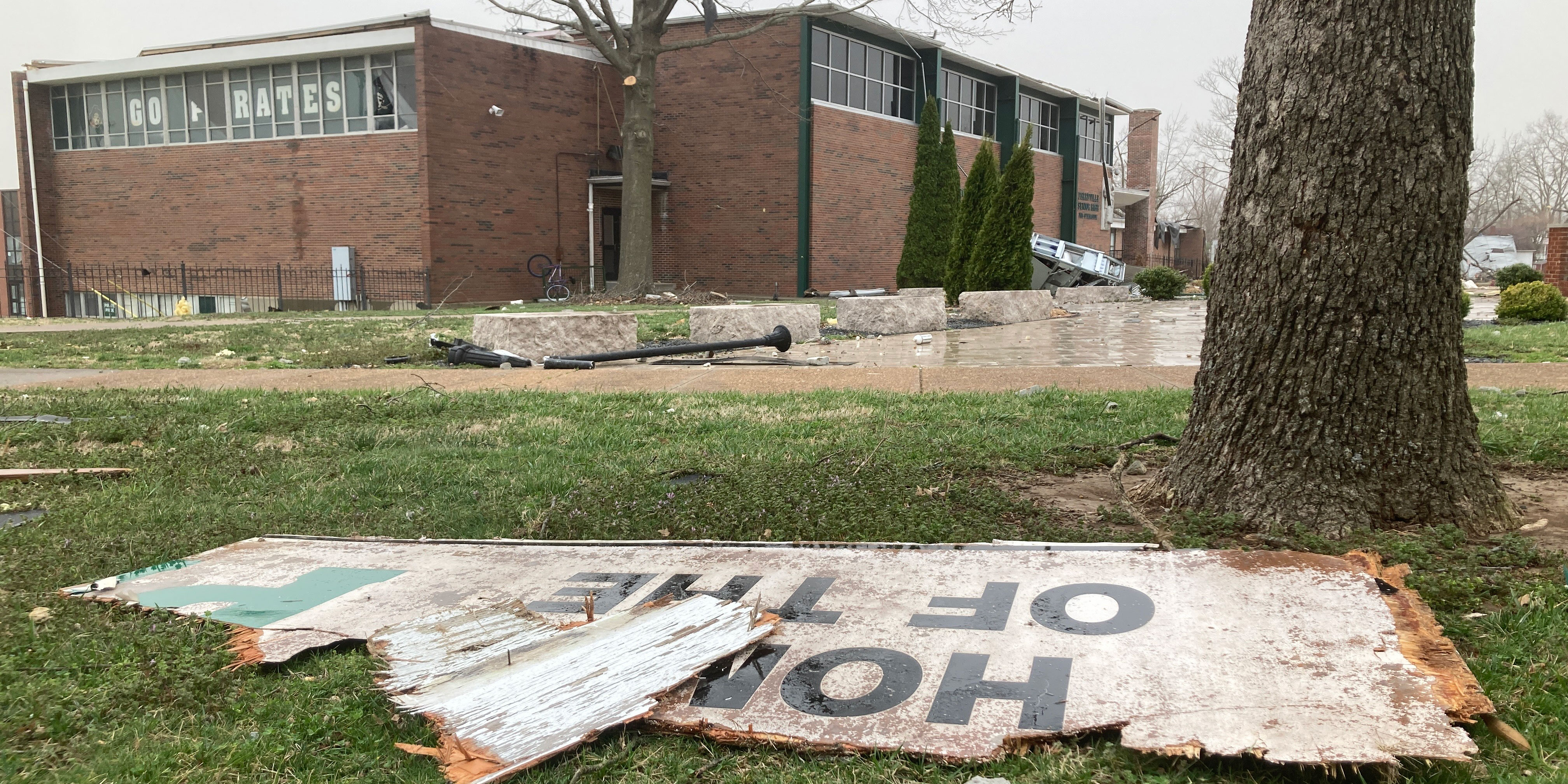 The damaged PHS building is in the background. Broken pieces of a sign lie on the ground in the foreground. The broken sign says "Home of the P.."