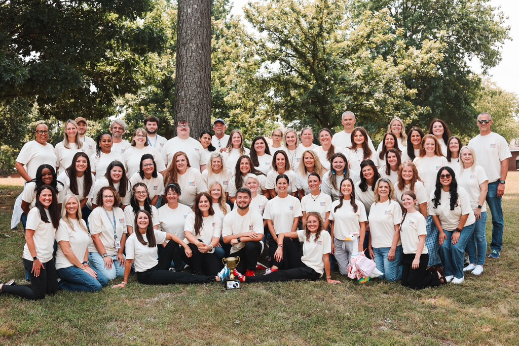 Group photo in a park; many people in white shirts, some with hats, posing under a large tree.