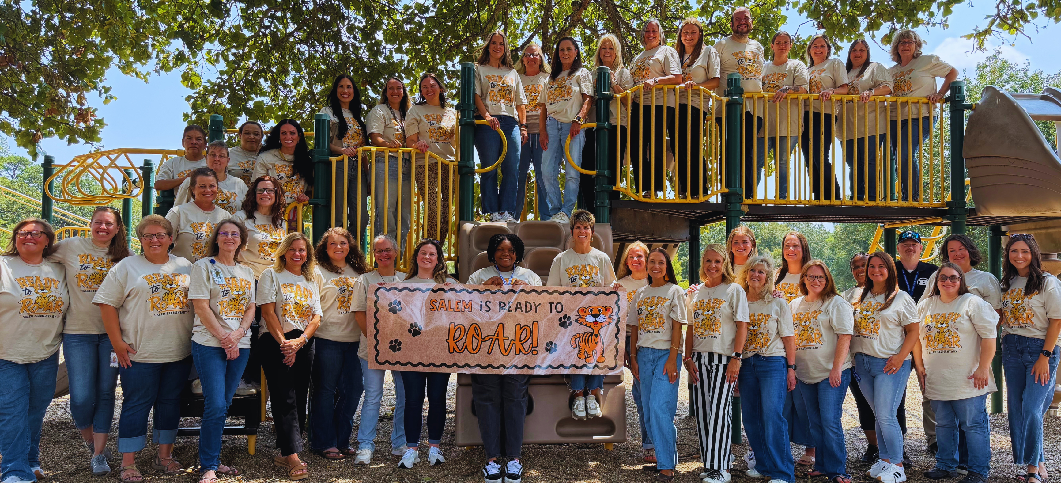 Large group of adults wearing matching T-shirts pose in front of a playground with a banner.