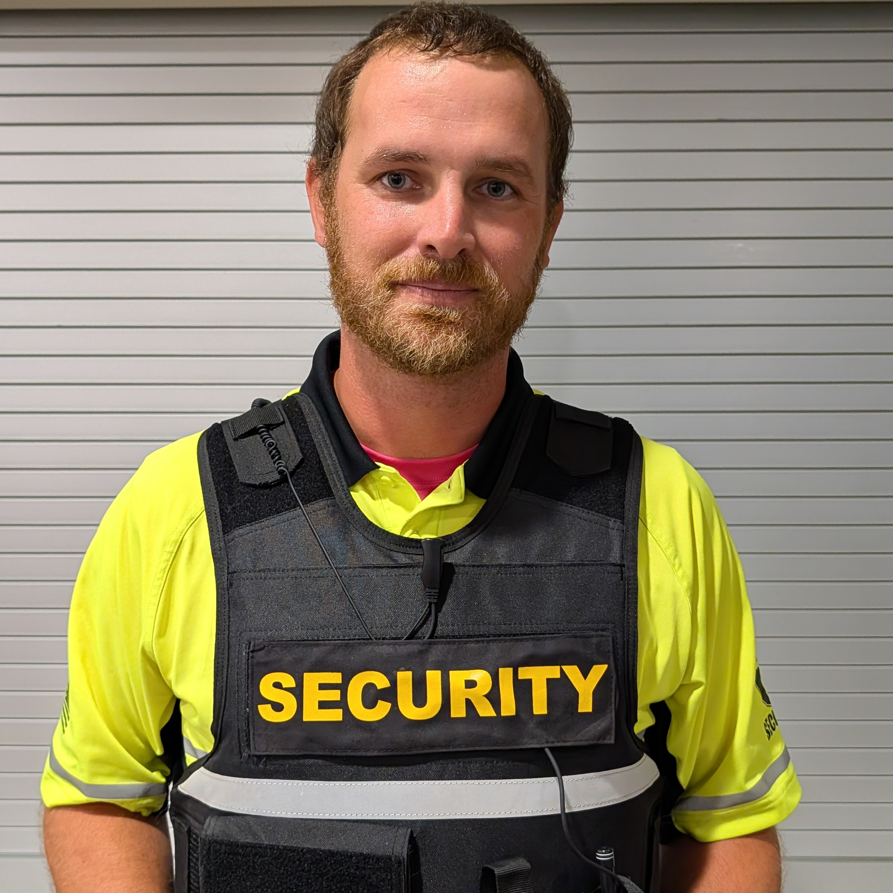 A security officer wearing a bright yellow shirt and black protective vest labeled “SECURITY” stands indoors in front of a gray background.