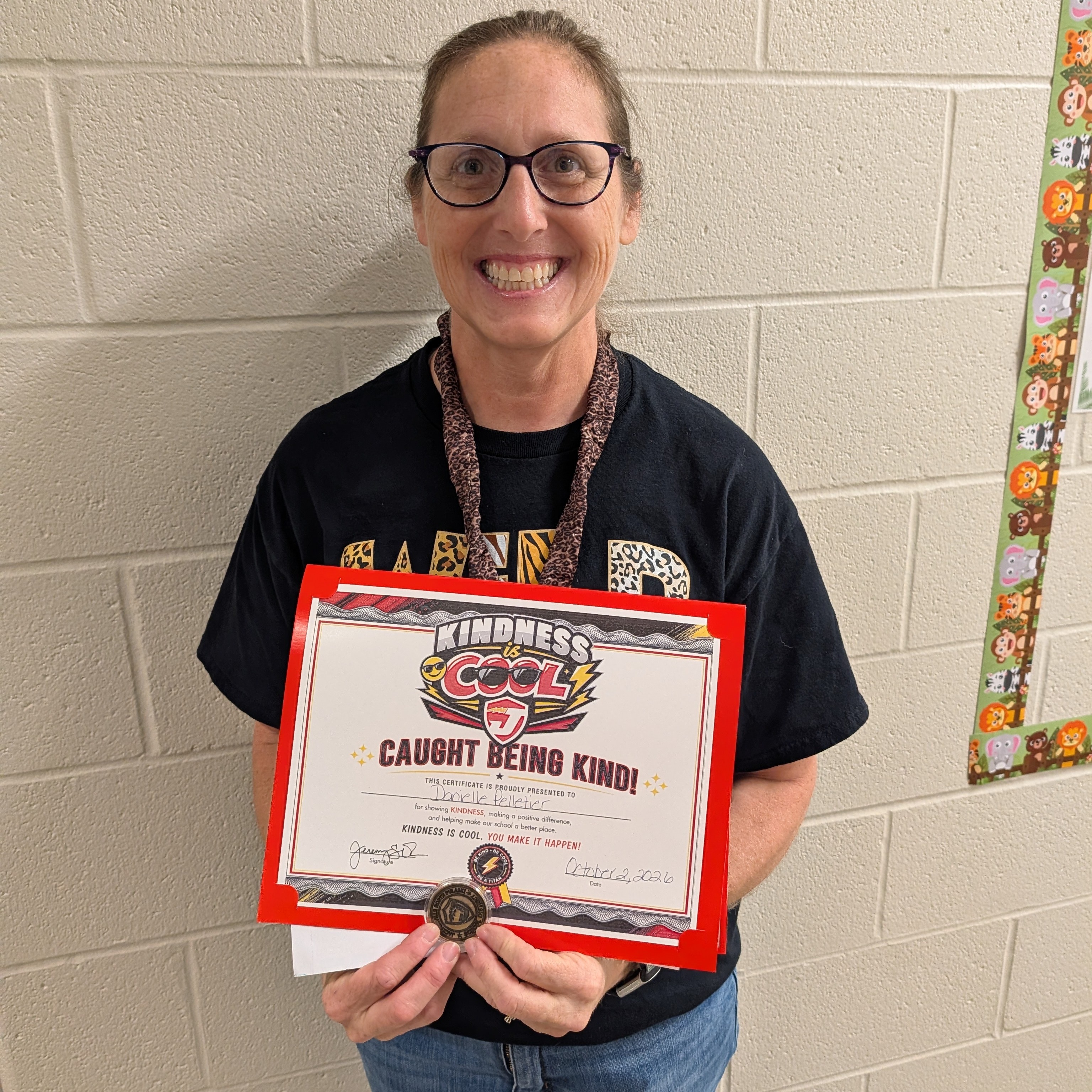 A smiling educator wearing glasses, a black shirt, and jeans stands in a school hallway holding a red-framed “Kindness Is Cool – Caught Being Kind!” certificate and recognition medal.