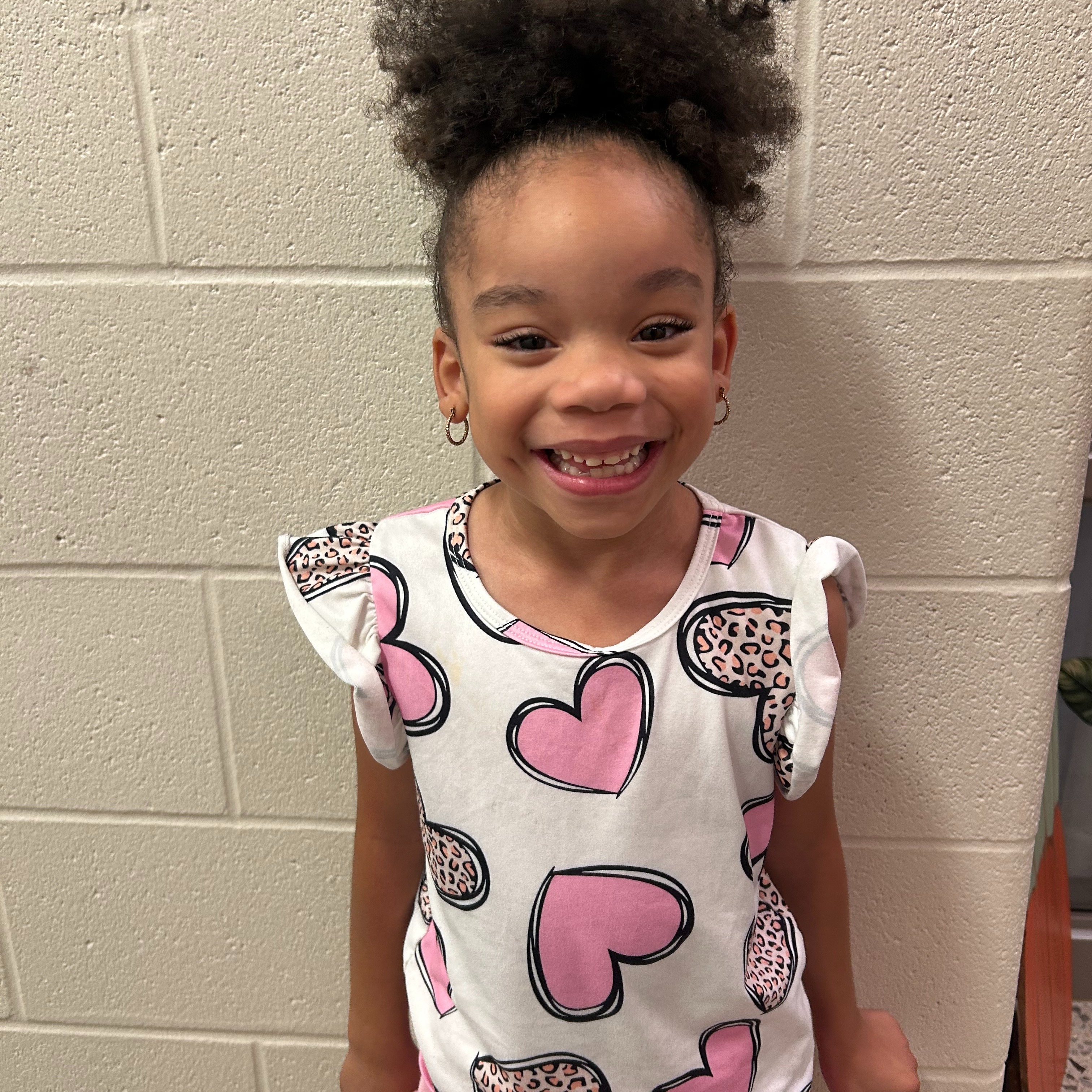 A smiling young scholar wearing a white shirt decorated with pink and leopard-print hearts and a pink ruffled skirt stands in front of a light-colored brick wall.