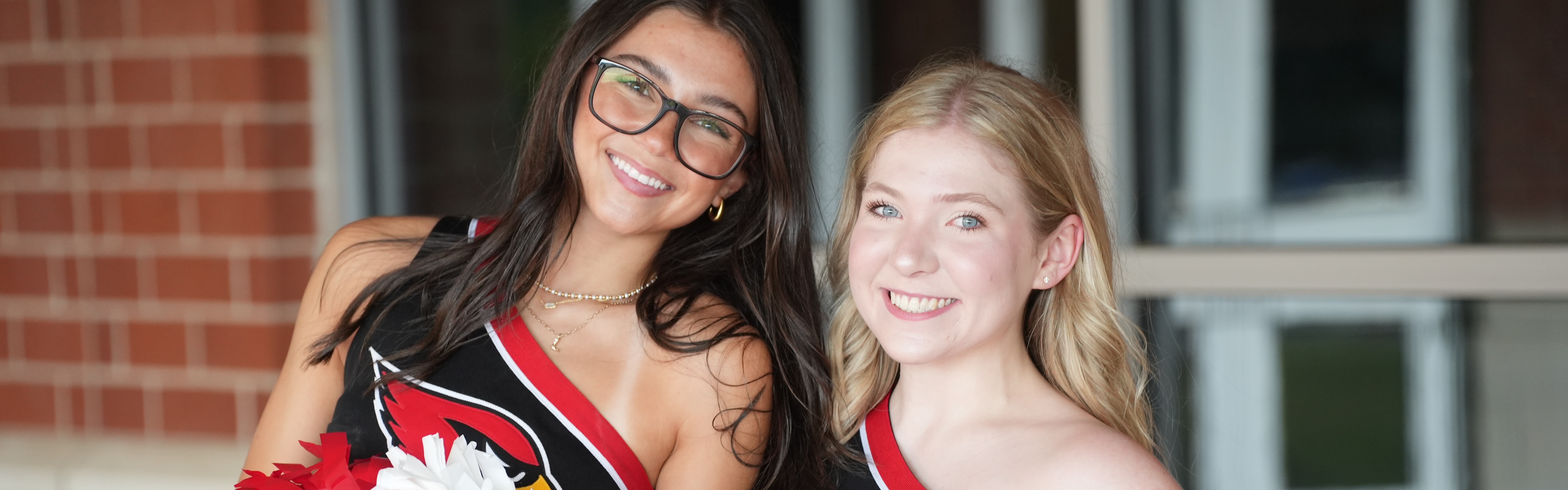 Two women in cheerleader uniforms, holding red and white pompoms, stand next to each other in front of a brick wall.