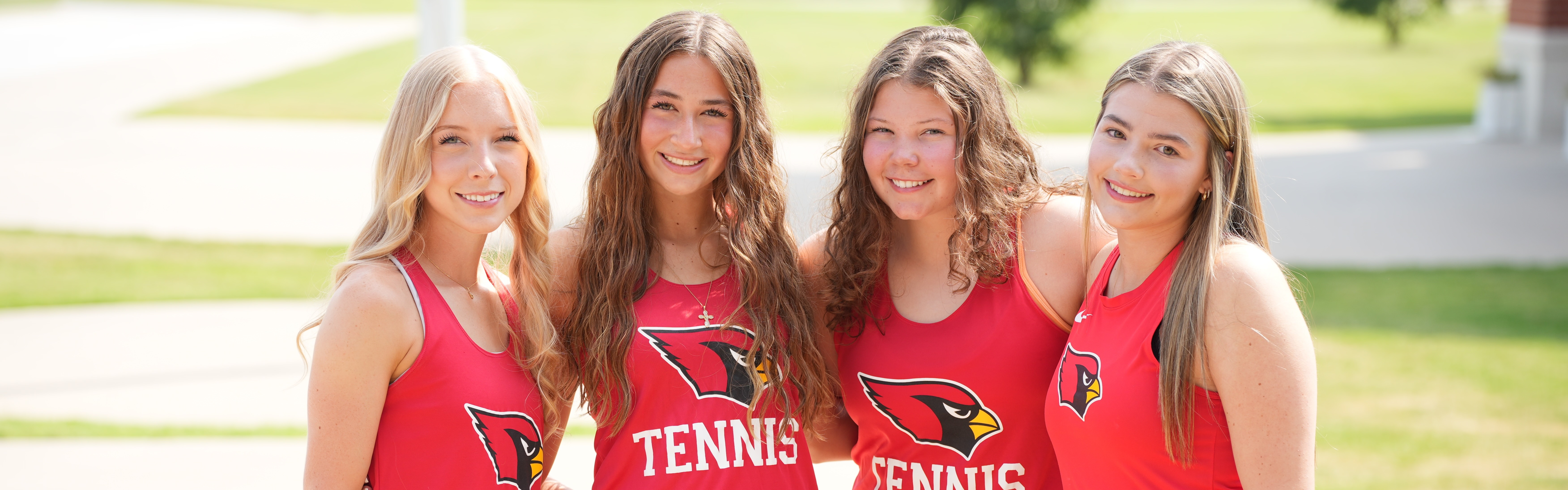 Four women wearing red tennis shirts and black skirts stand close together on a sidewalk. Behind them is a grassy area.
