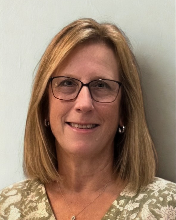Woman with short blonde hair wearing glasses, a floral top, and a necklace. She is smiling.