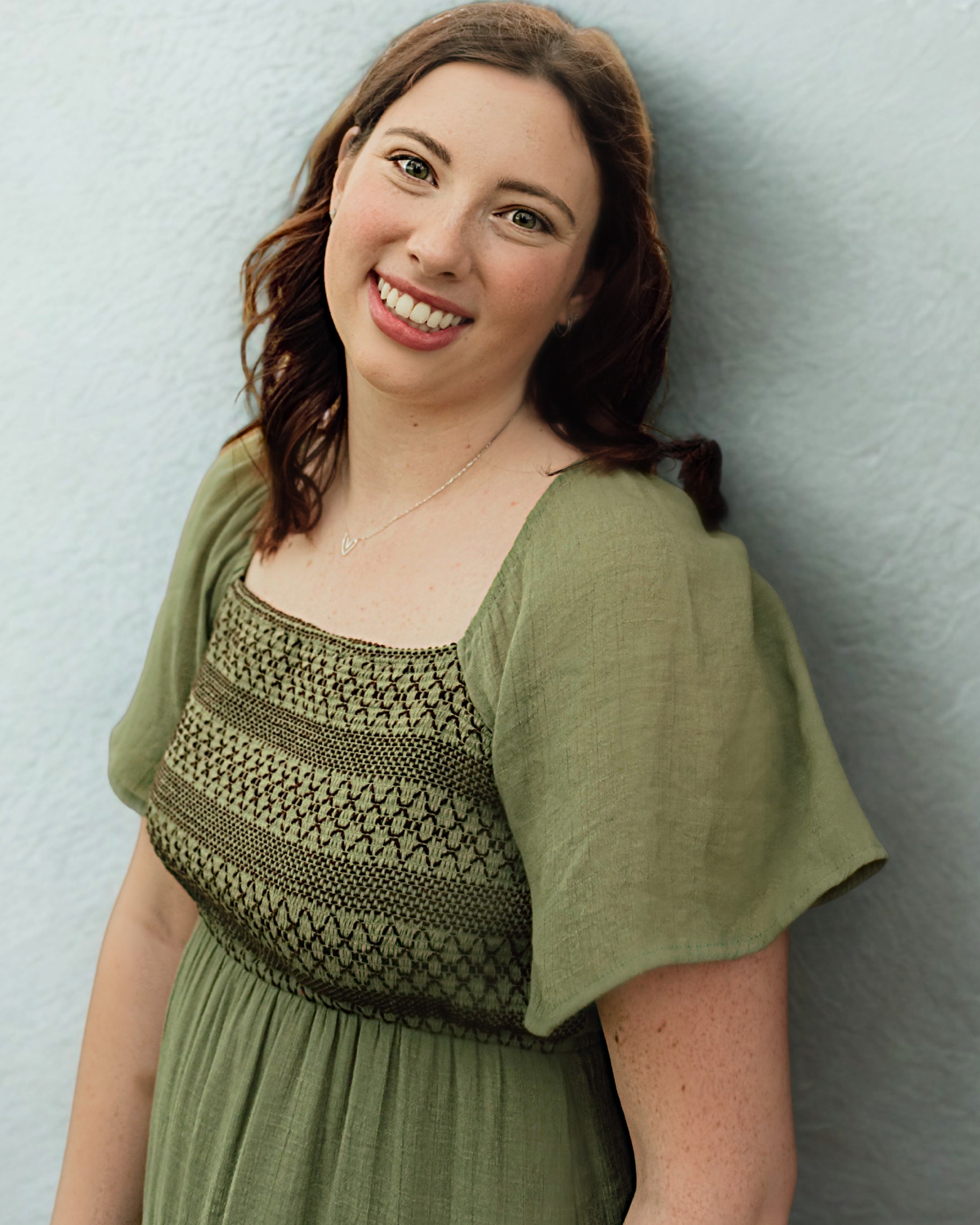A woman smiling, wearing a green dress with intricate patterns. She has brown hair and is against a light blue wall.