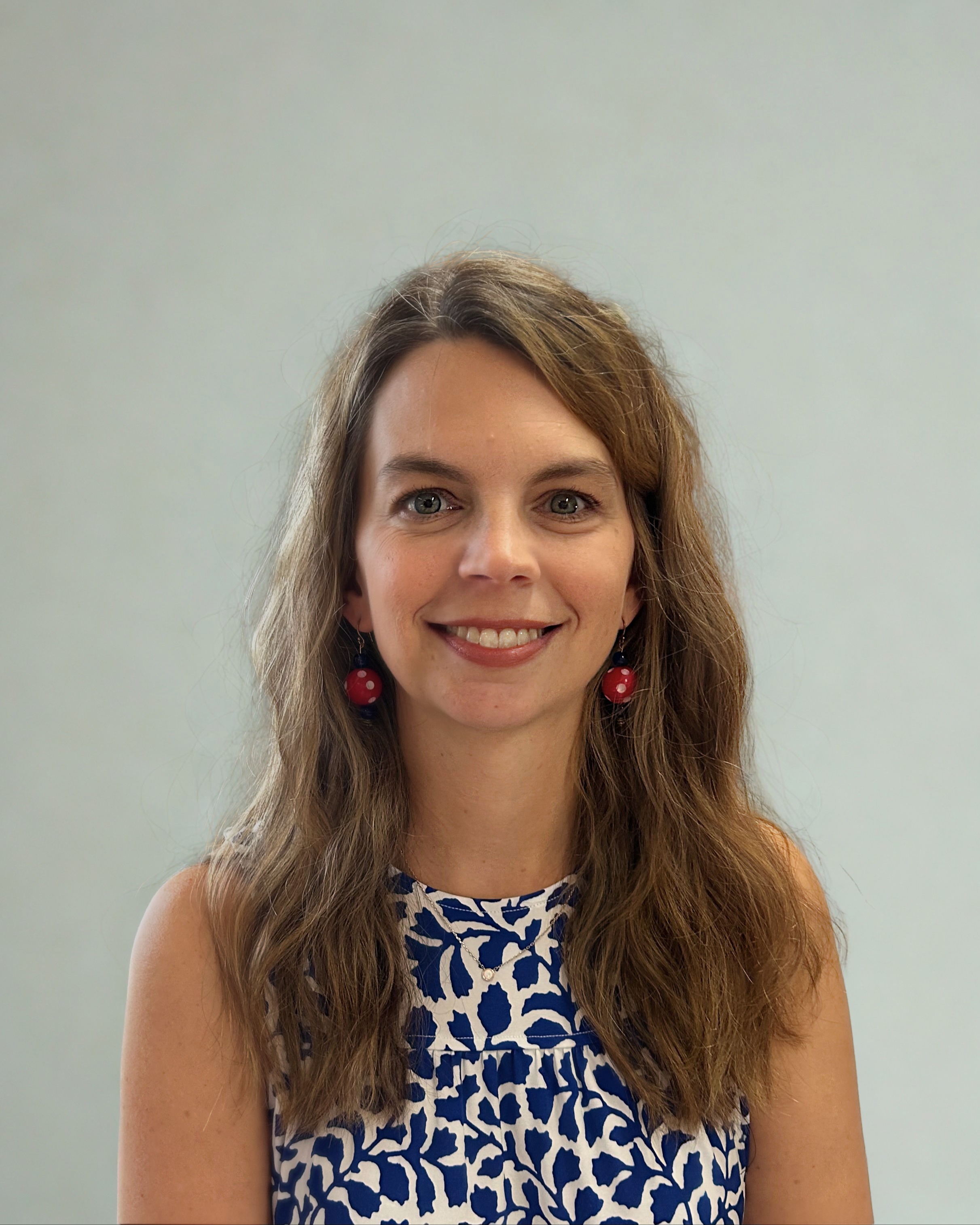 Woman with long wavy brown hair, wearing blue and white patterned top, red earrings, and smiling at camera.