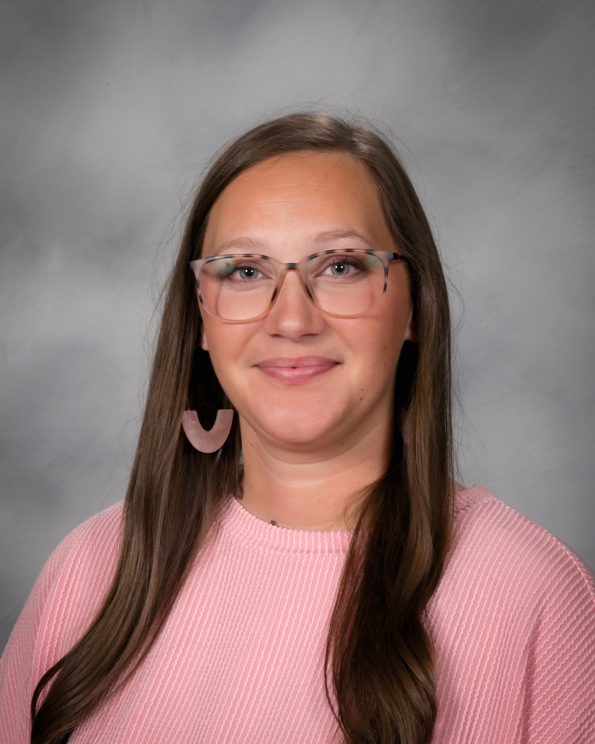 Adult woman wearing glasses and a pink sweater, with long hair, standing against a gray background.