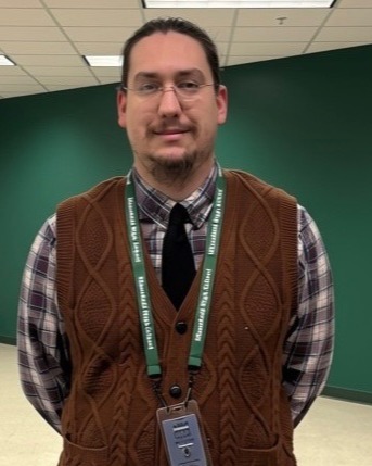 Man in a plaid shirt wearing a brown cardigan, glasses, tie, and ID. Standing in a hallway with green wall and checkerboard floor.