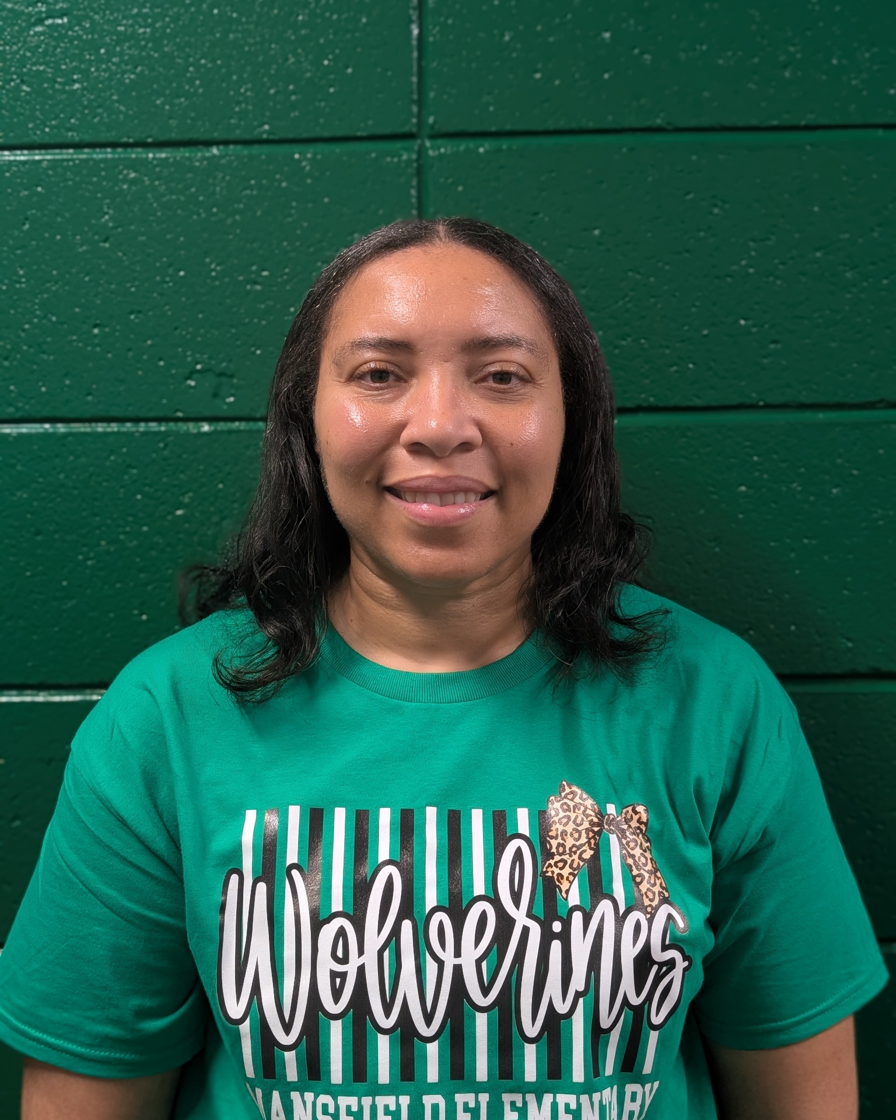 Adult woman standing against green wall wearing a green t-shirt with "Wolverines" and "Hansfield Elementary" printed on it.