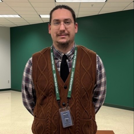 A man wearing glasses, a brown vest, plaid shirt, black tie, and ID lanyard stands in a room with green walls and checkered floor.