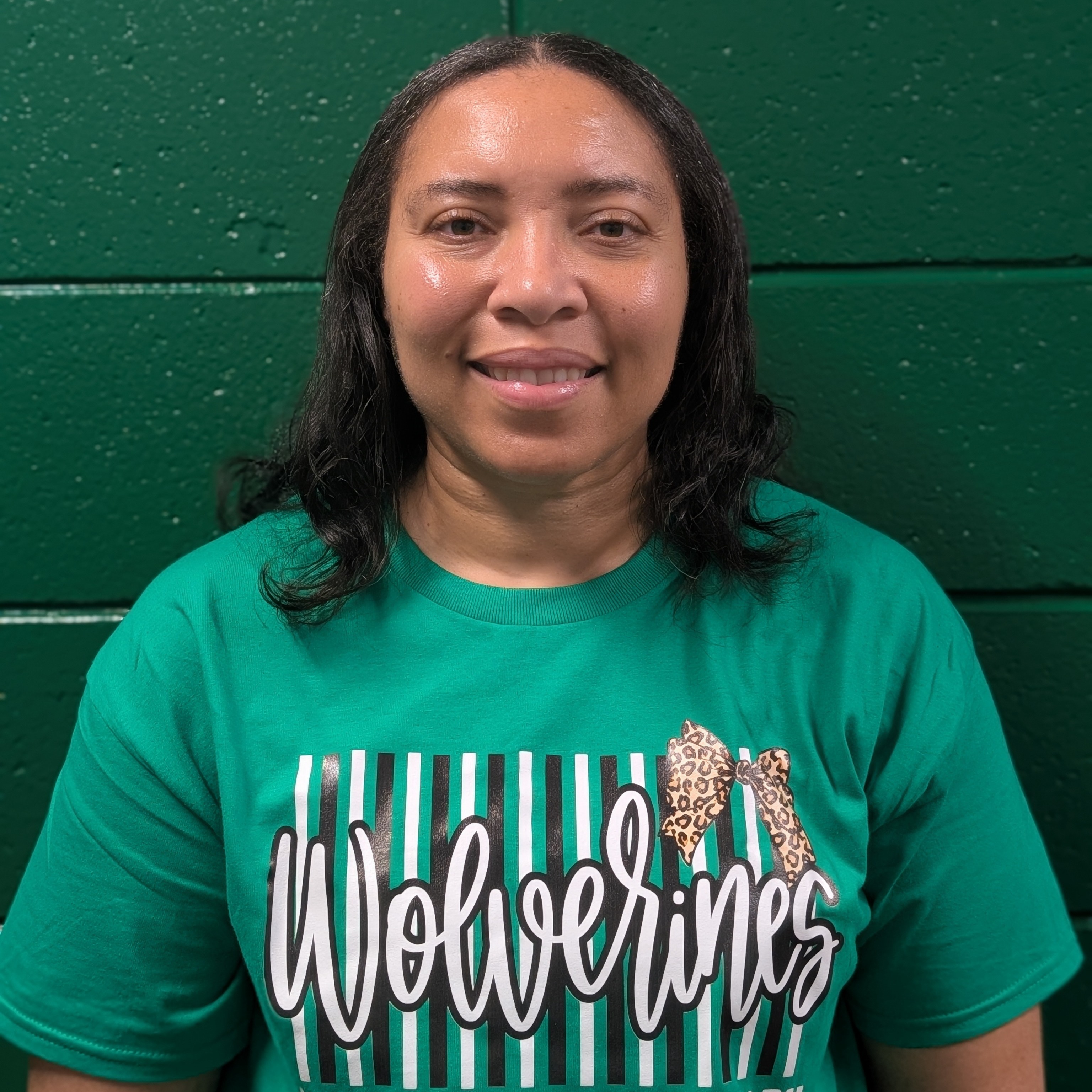 Woman wearing green T-shirt with Wolverines logo. Standing in front of green wall.
