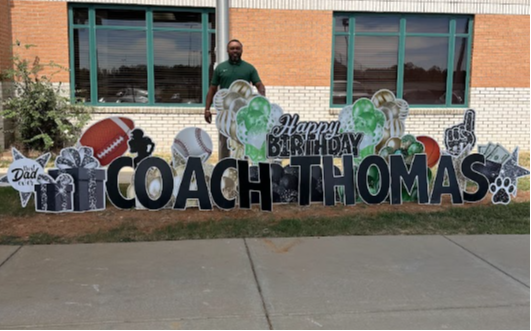 Person stands near a birthday sign with text, footballs, and other sports items. Building with windows in the background.